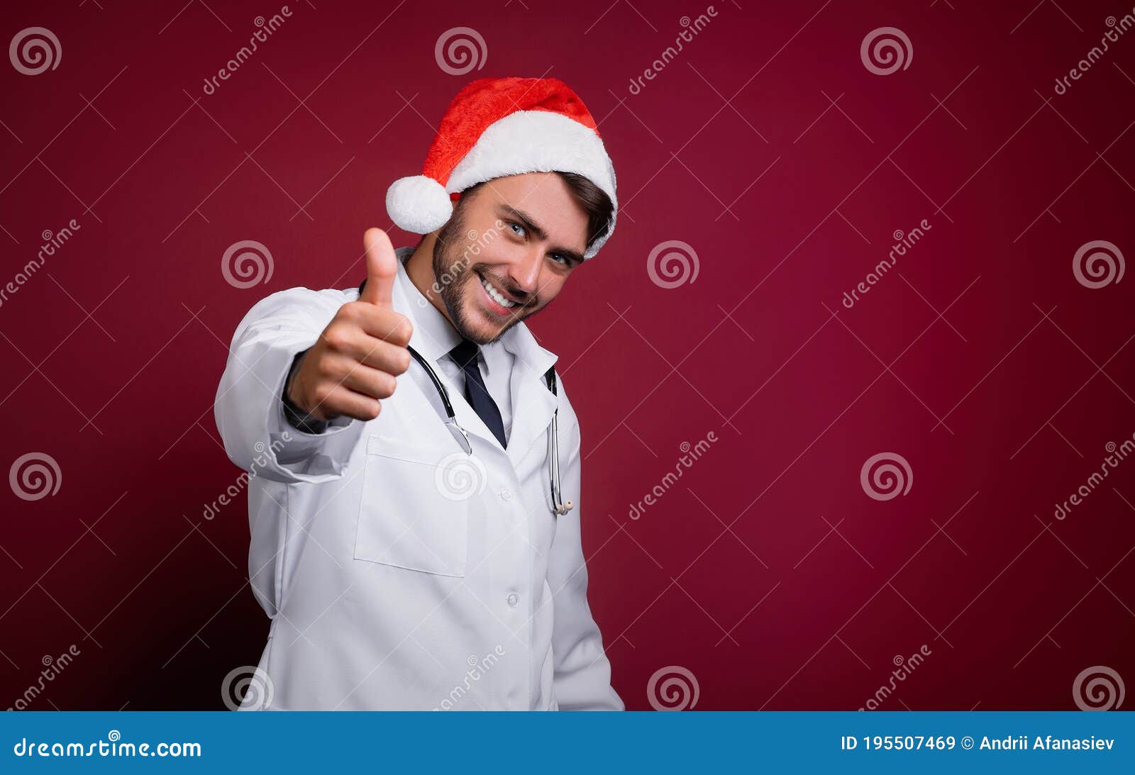 young handsome doctor in white uniforme and santa claus hat standing in studio on red background smile and finger in camera