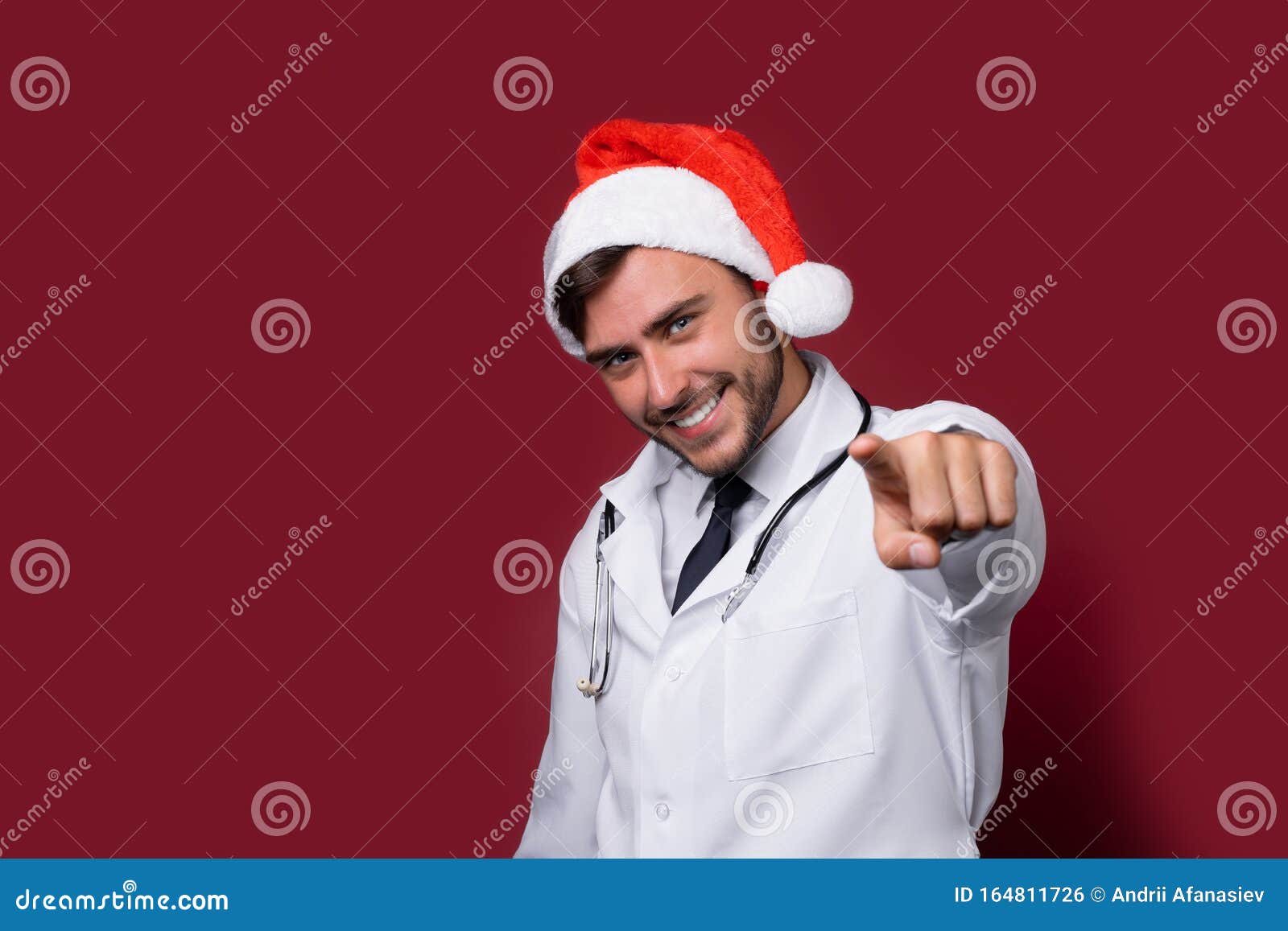 young handsome doctor in white uniforme and santa claus hat standing in studio on red background smile and finger in camera