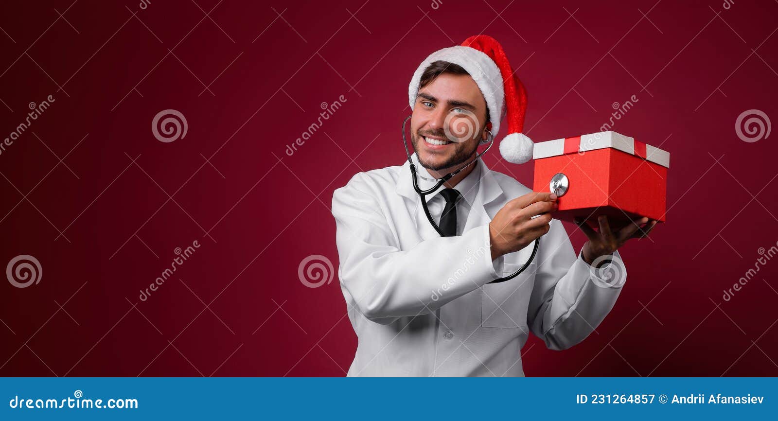 young handsome doctor in white uniforme and santa claus hat standing in studio on red background smile and finger in camera