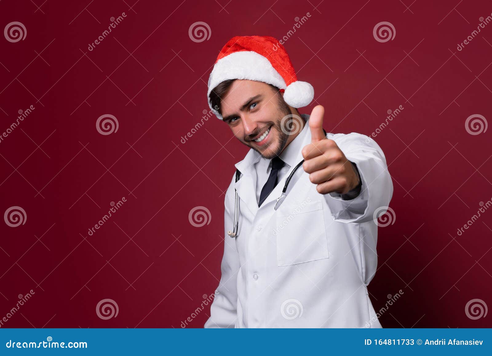 young handsome doctor in white uniforme and santa claus hat standing in studio on red background smile and showing a thumbs up