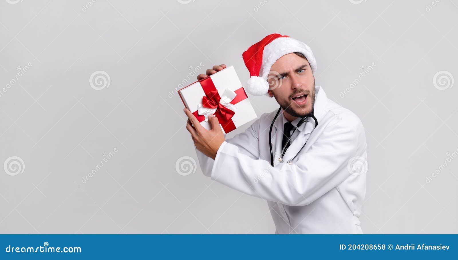 young handsome doctor in white uniforme and santa claus hat standing in studio on white background smile and finger in camera