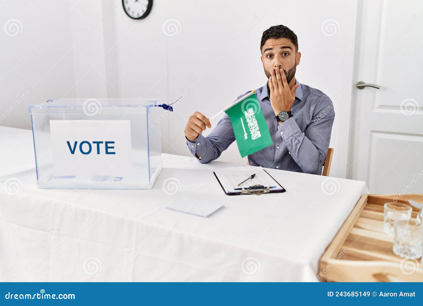 young handsome man with beard at political campaign election holding arabia saudita flag covering mouth with hand, shocked and