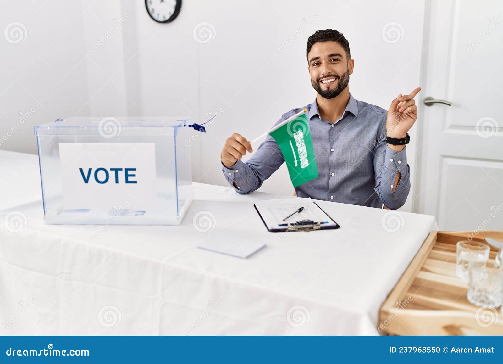 young handsome man with beard at political campaign election holding arabia saudita flag smiling happy pointing with hand and