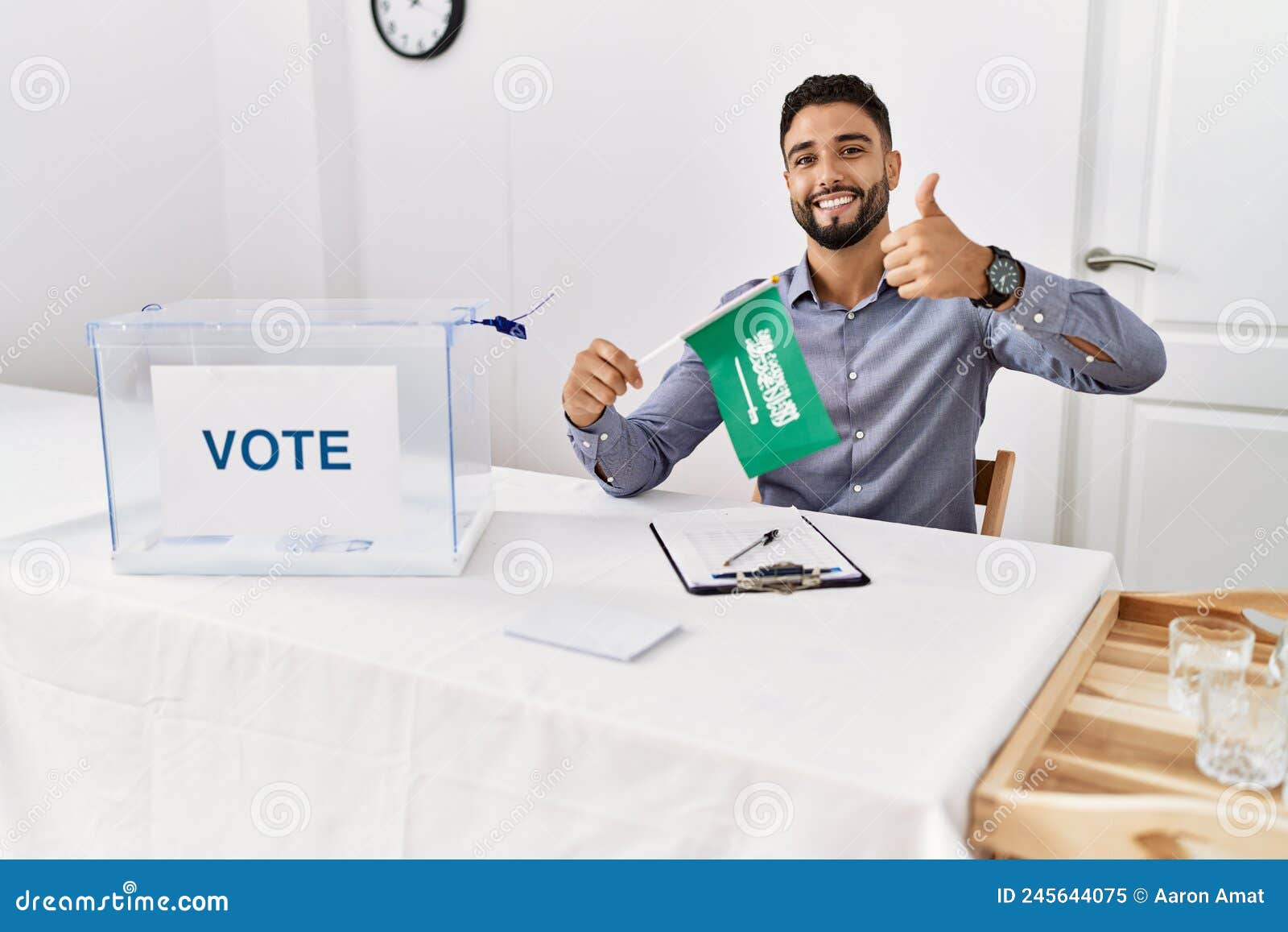 young handsome man with beard at political campaign election holding arabia saudita flag smiling happy and positive, thumb up