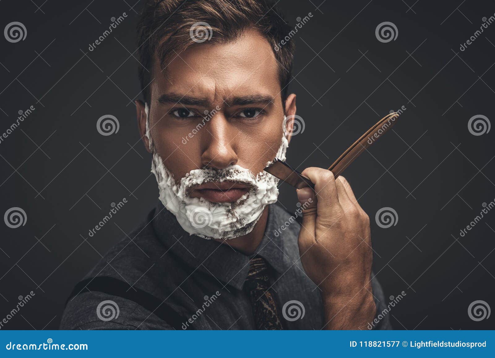 young handsome man with shaving cream on his face, grooming his beard with straight razor and looking