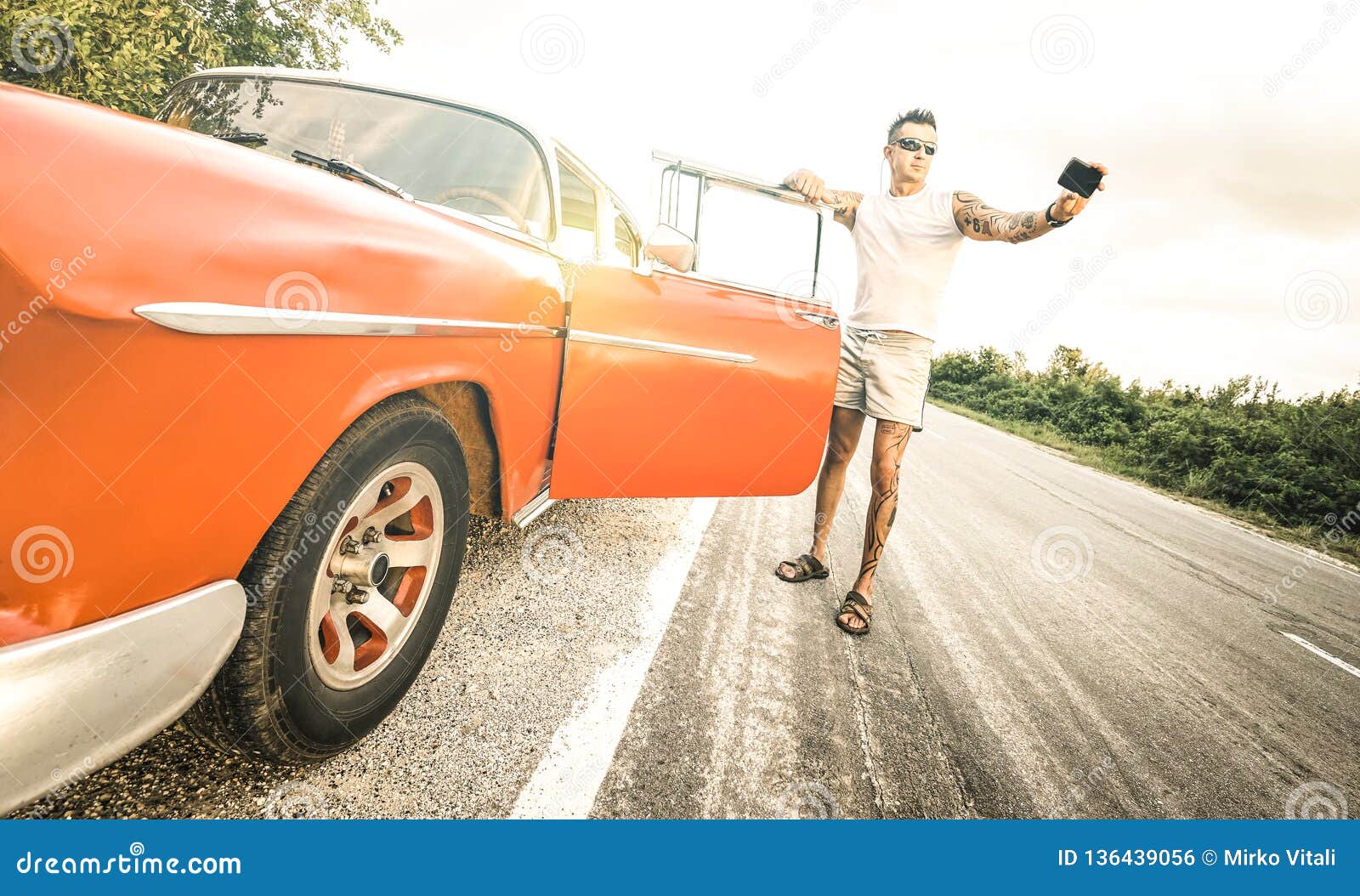 young hipster fashion man with tattoo taking selfie with vintage car during road trip in cuba - travel wanderlust concept
