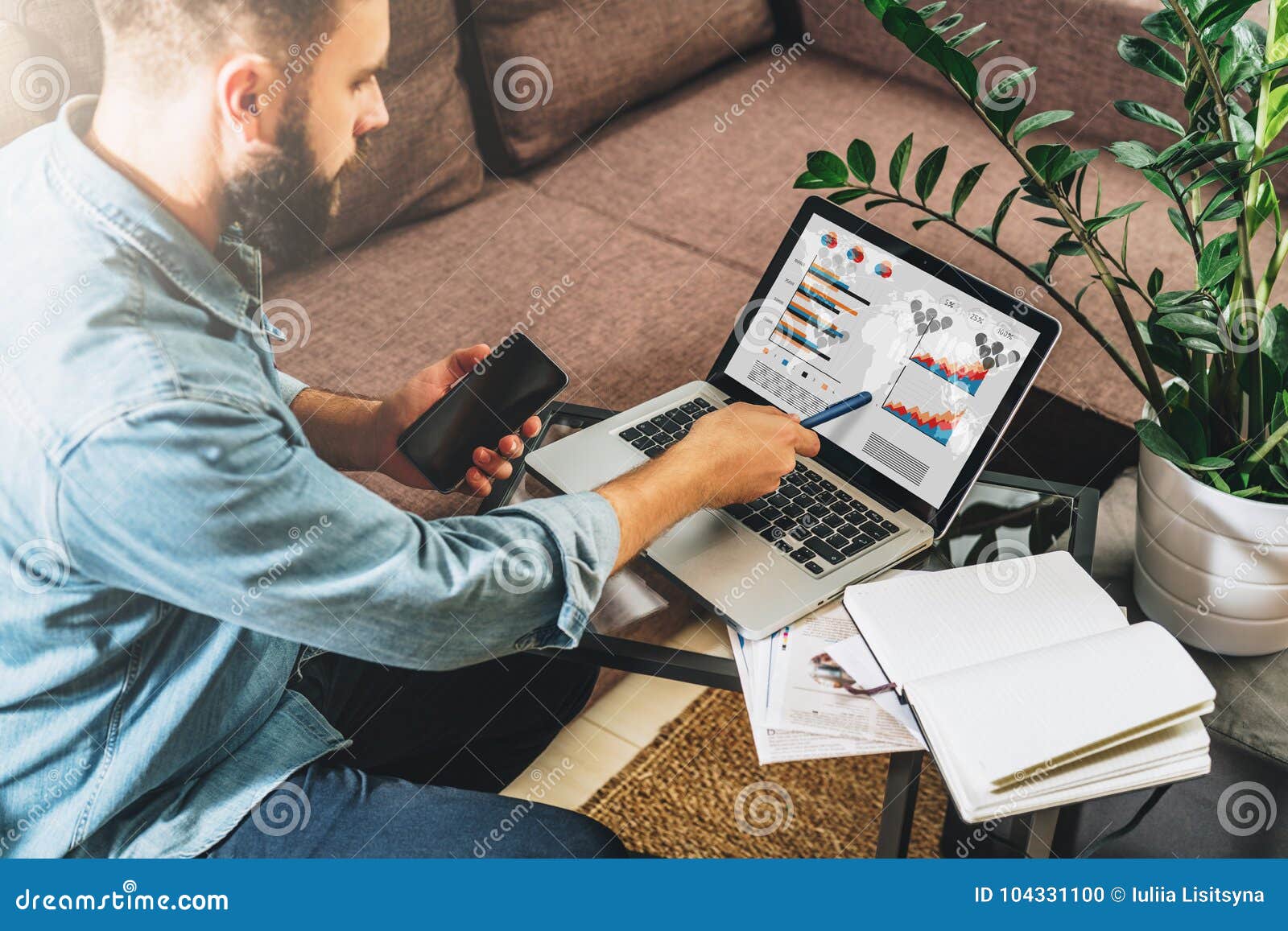 young hipster man, entrepreneur sits at home on couch at coffee table, holding smartphone, showing pencil on screen