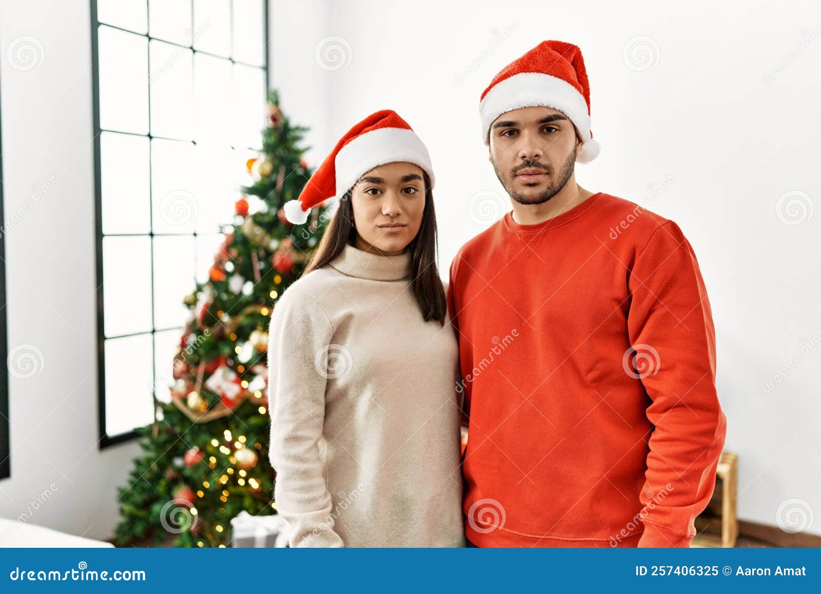 Young Hispanic Couple Standing by Christmas Tree Relaxed with Serious