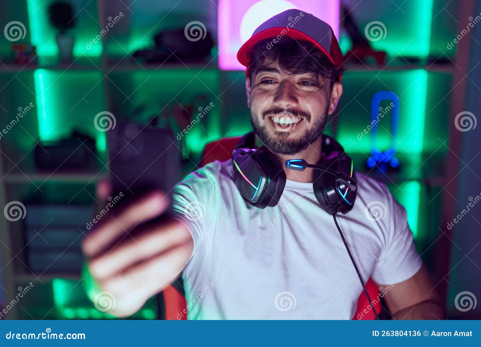 young hispanic man streamer using computer make selfie by smarpthone at gamin room