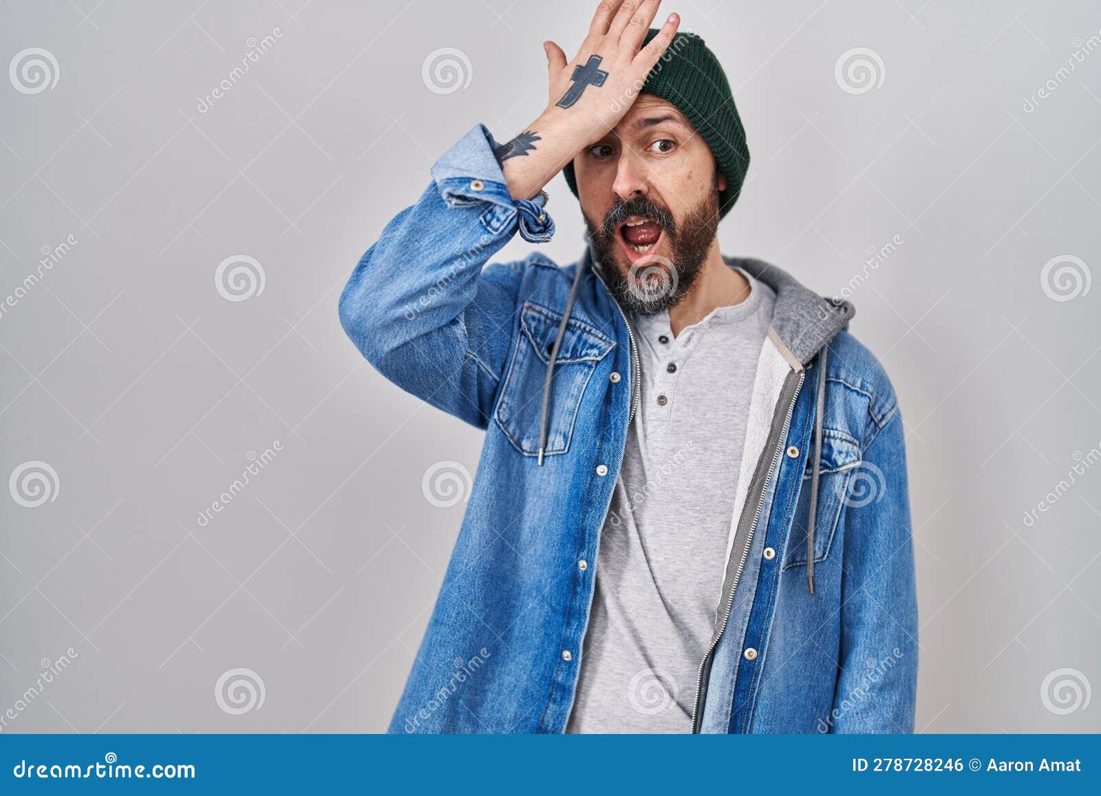 young hispanic man with tattoos wearing wool cap surprised with hand on head for mistake, remember error
