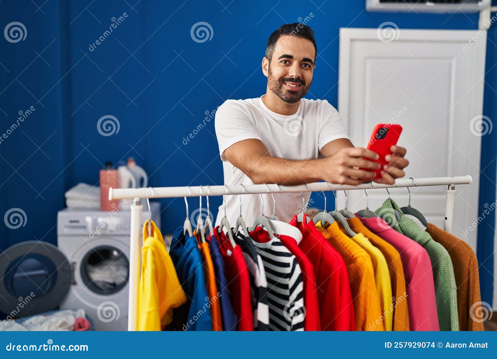 Young Hispanic Man Using Smartphone Leaning on Clothes Rack at Laundry