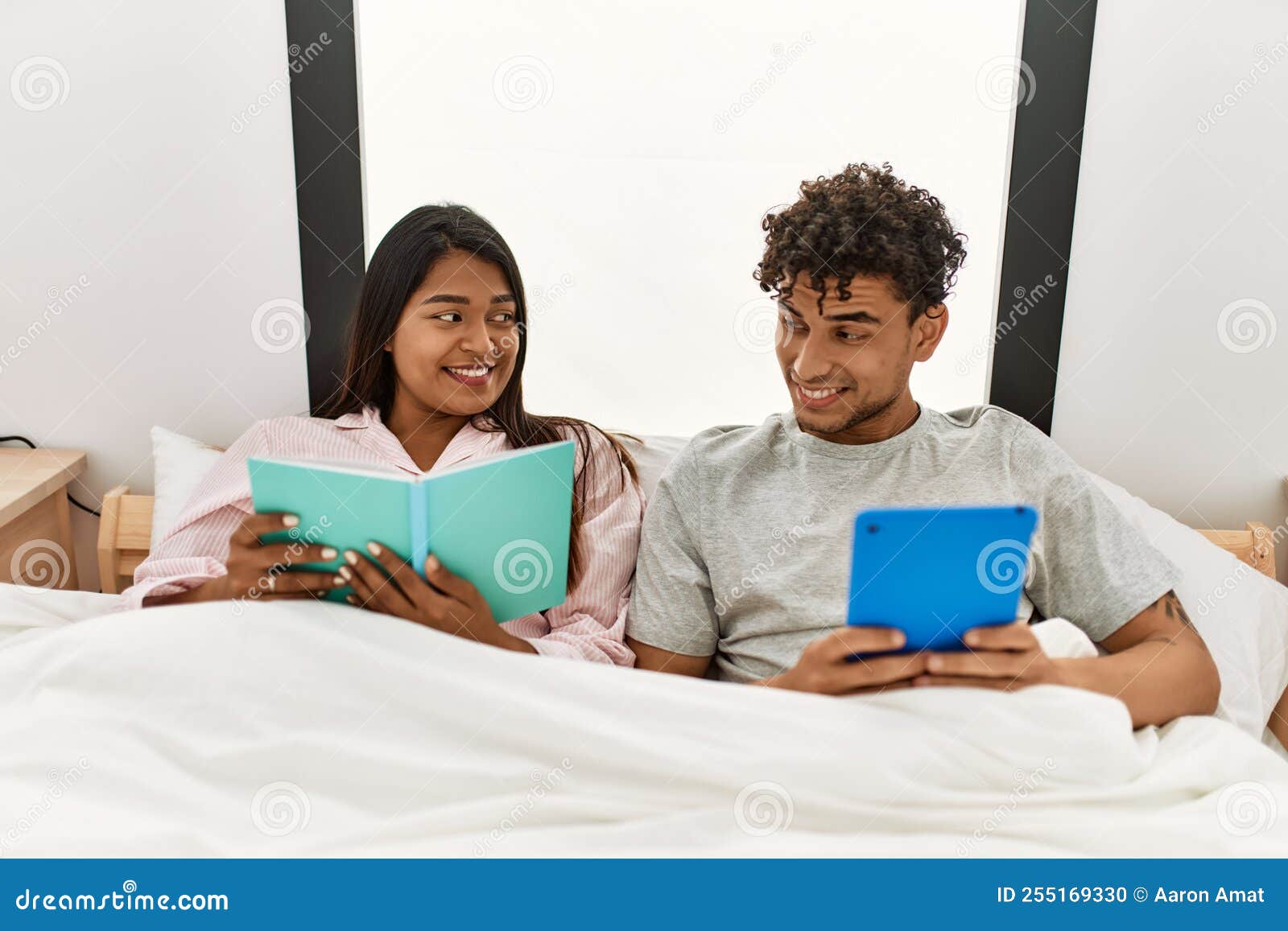 Young Latin Couple Reading Book Using Touchpad Sitting on the Bed at Bedroom Stock Photo Image