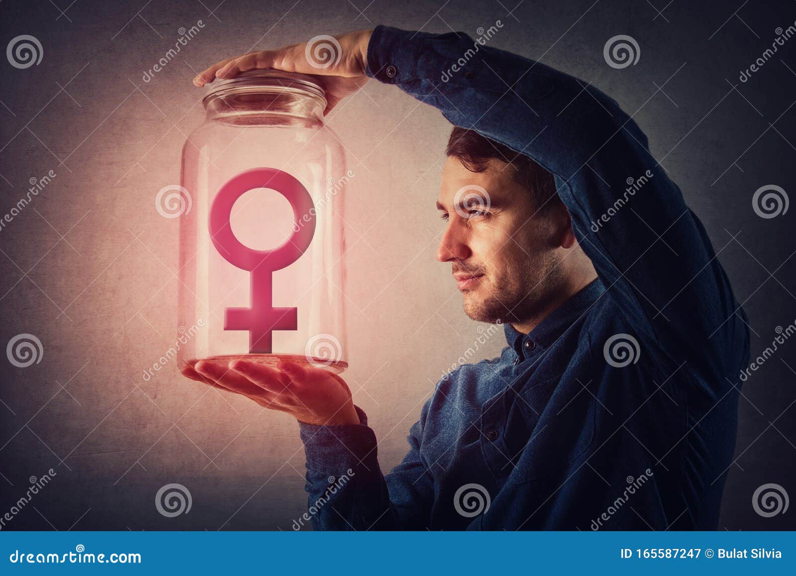 young male, malefic look holding a glass jar with female gender  inside as captive. man pretending to be superior to woman,