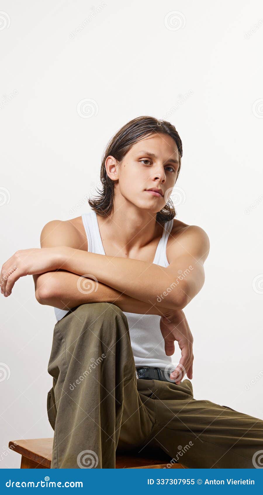 young man dressed white t-shirt and hacky color pants posing confidently, sitting on wooden stool against white studio