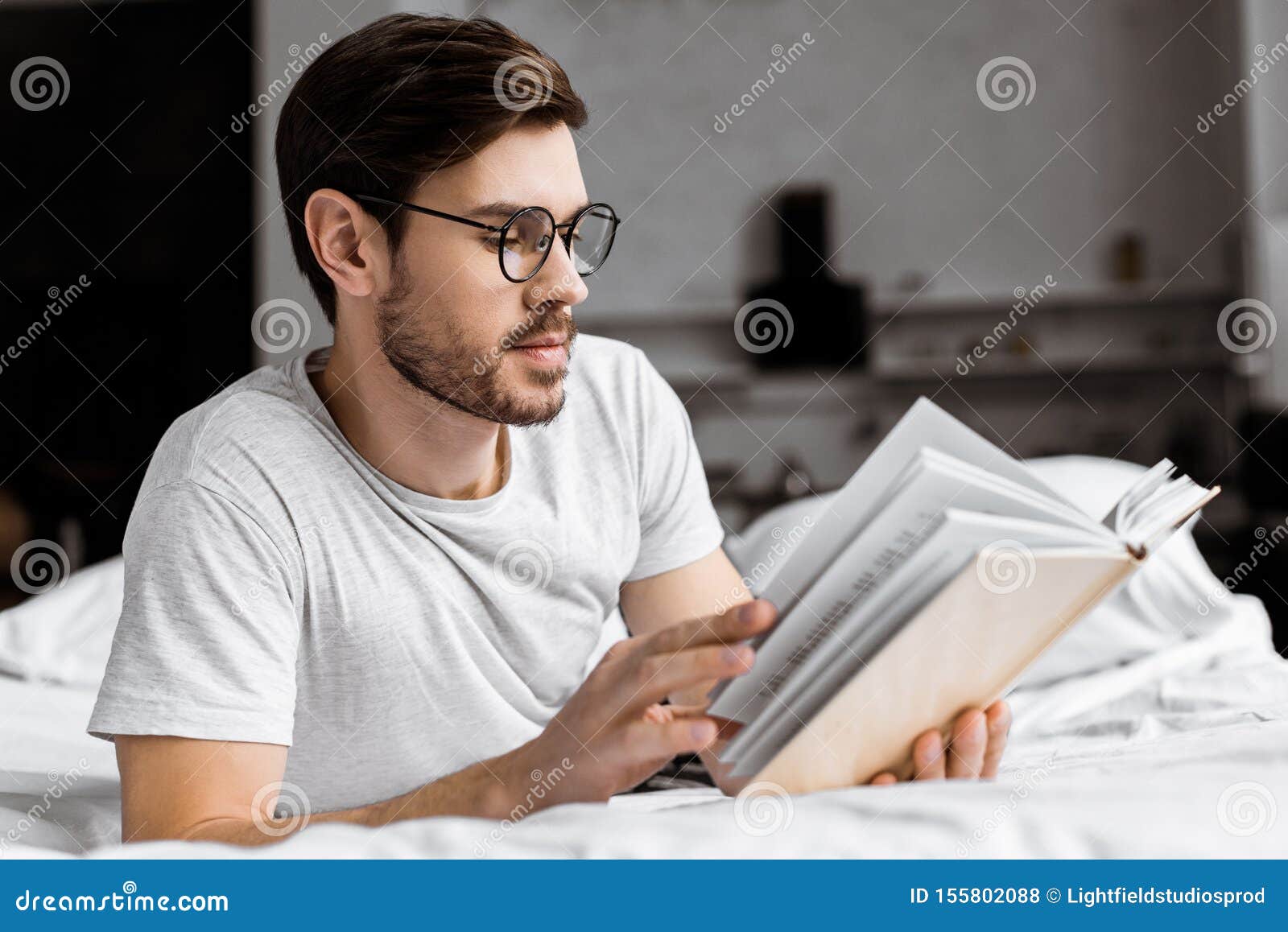 Young Man in Eyeglasses Lying in Bed and Reading Book Stock Photo
