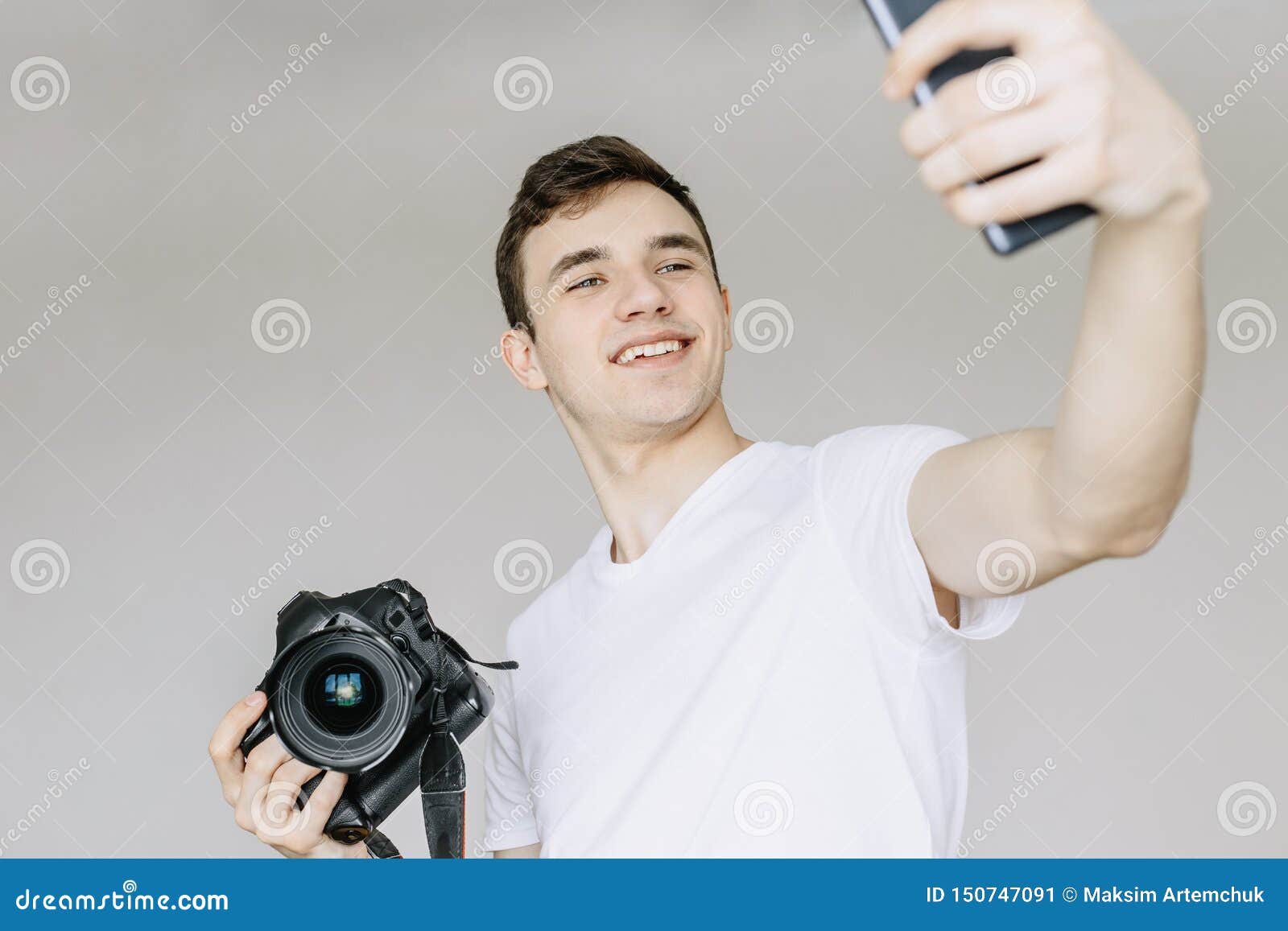 a young man holds a photo camera in his hand and makes a selfie by phone.  gray background