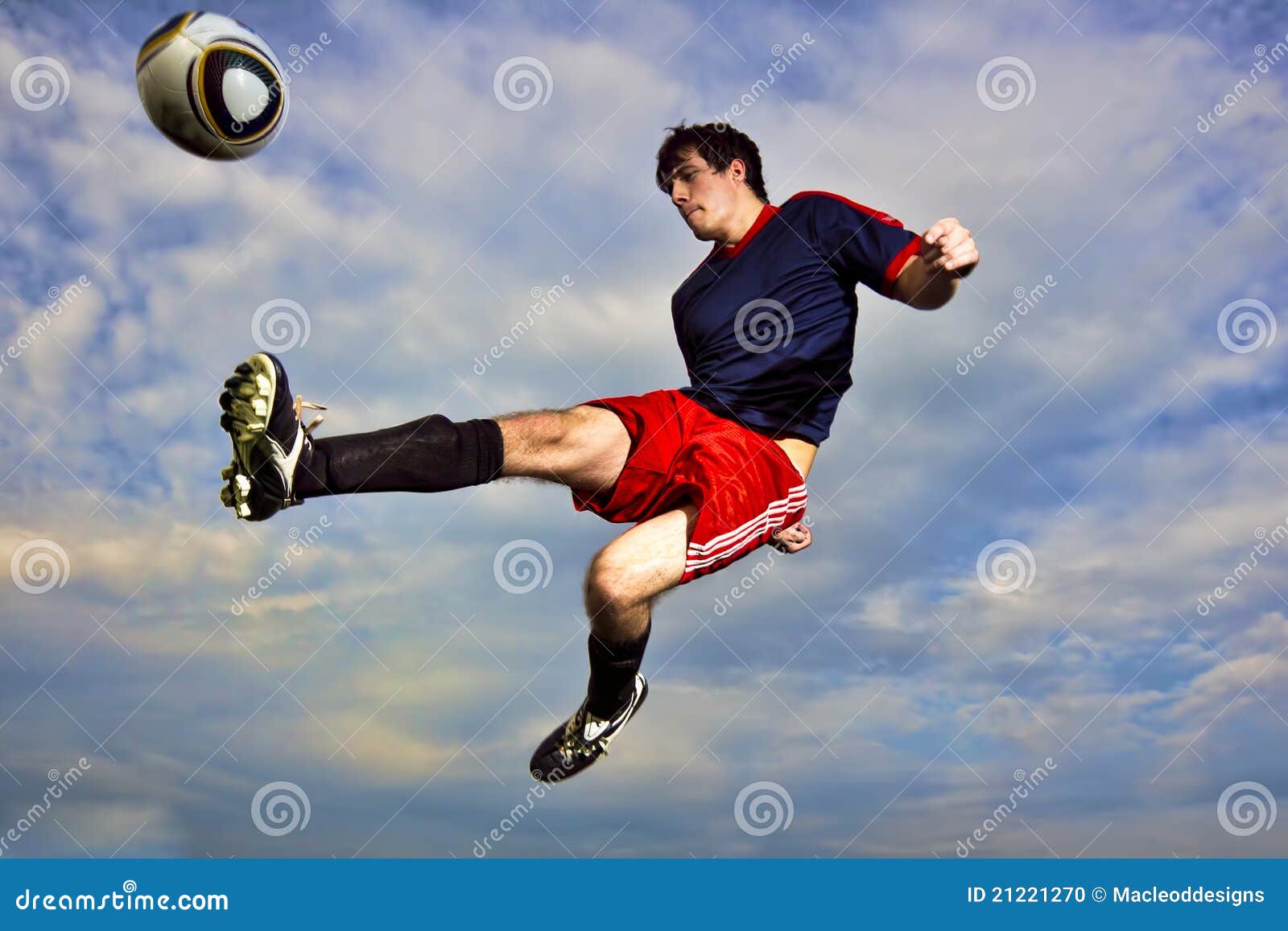 A young man kicks a soccerball midair. A young man in the air against a blue cloudy sky kicking a soccer ball