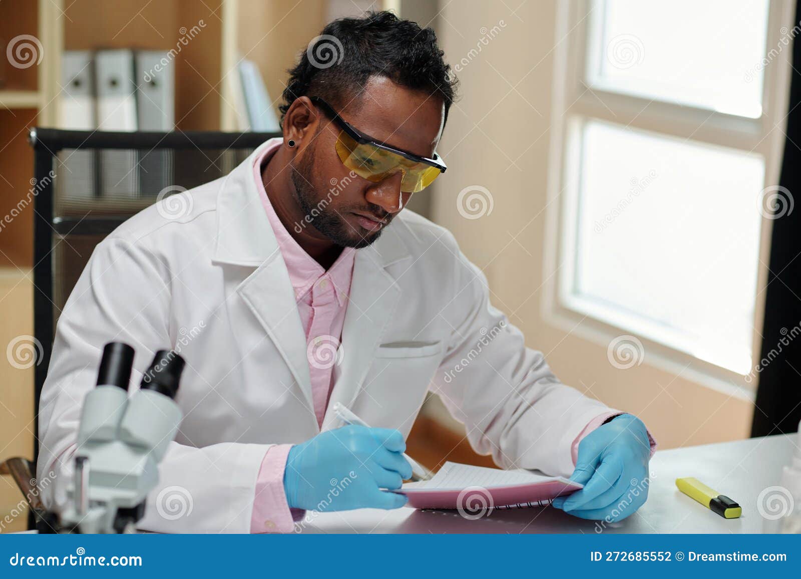 Young Man in Lab Coat and Gloves Making Notes in Copybook Stock Photo
