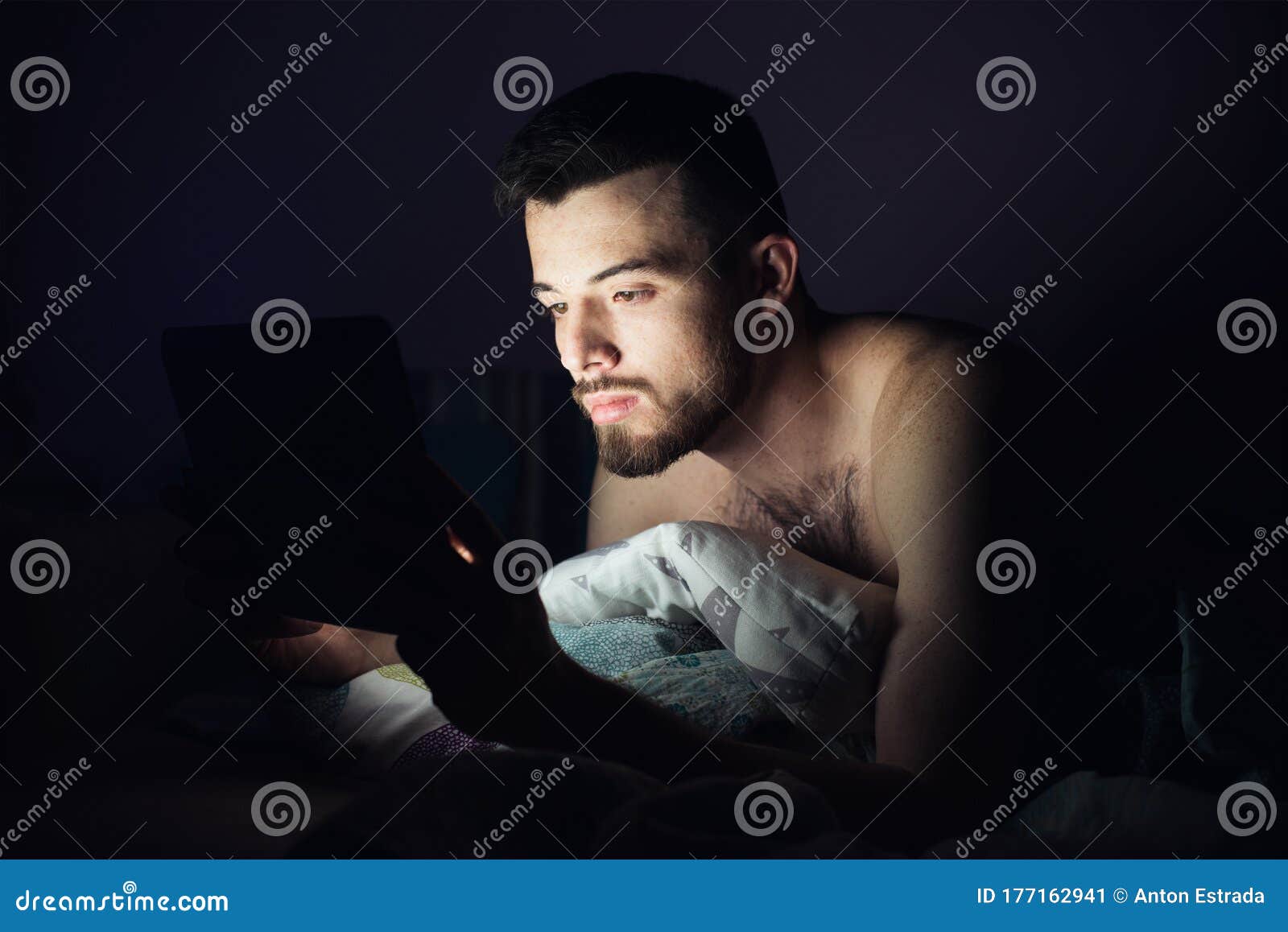 young man lying on bed on his belly and look at tablet`s screen. concentrated calm guy working playing games or