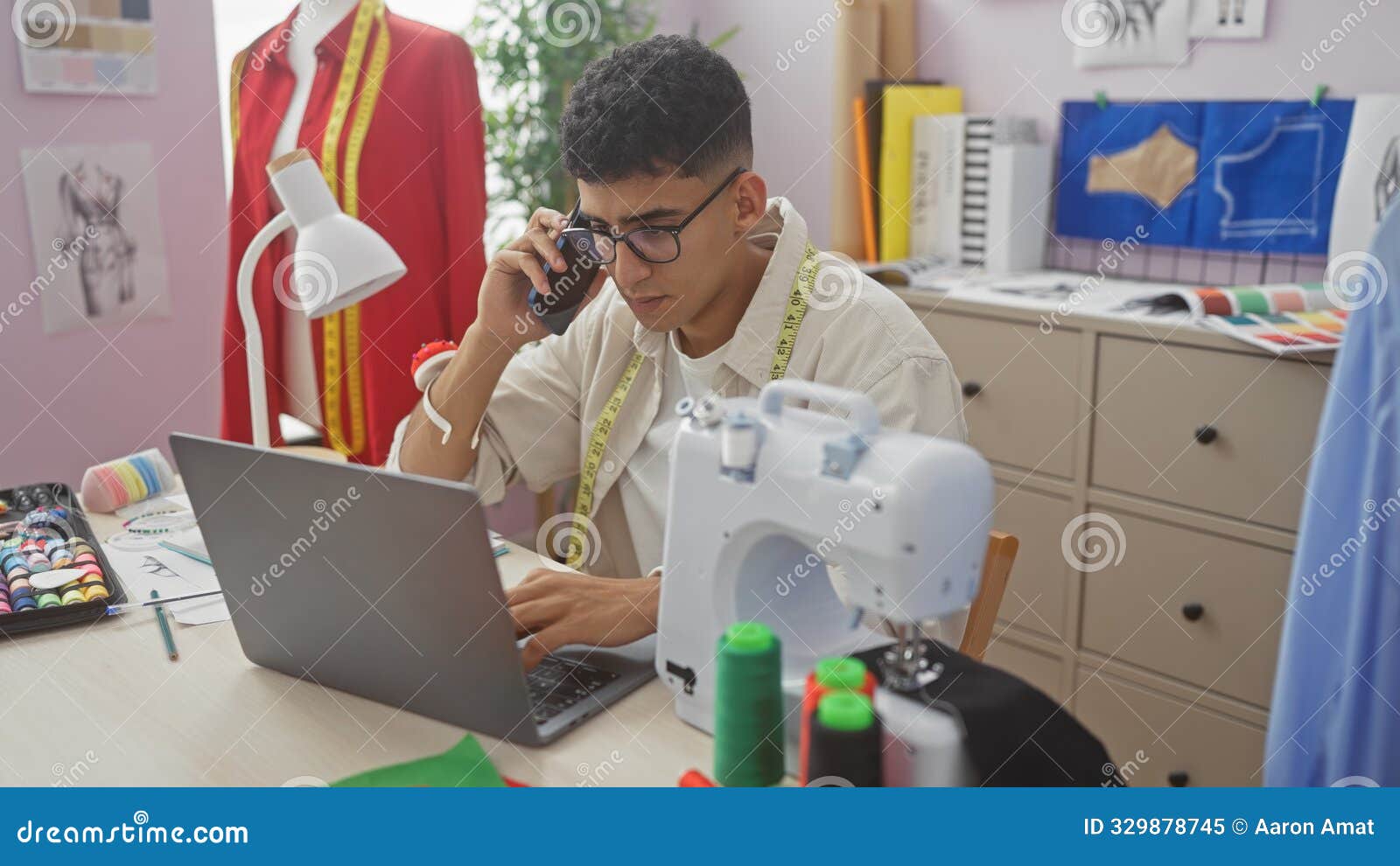 a young man multitasks at a tailor shop, using a phone and laptop amidst sewing equipment and garment drafts