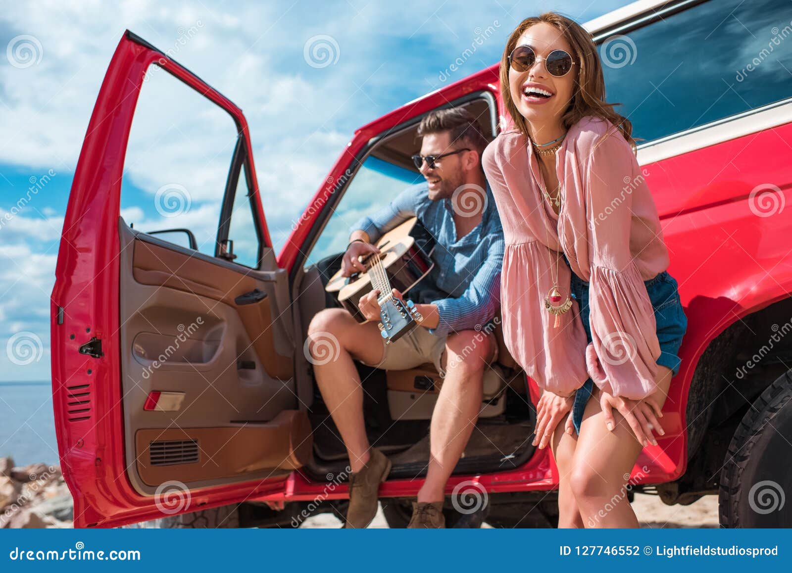 young man playing guitar near car with cheerful girlfriend