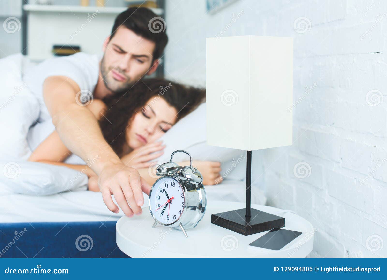 Young Man Reaching For Alarm Clock While Sleeping With Girlfriend Stock