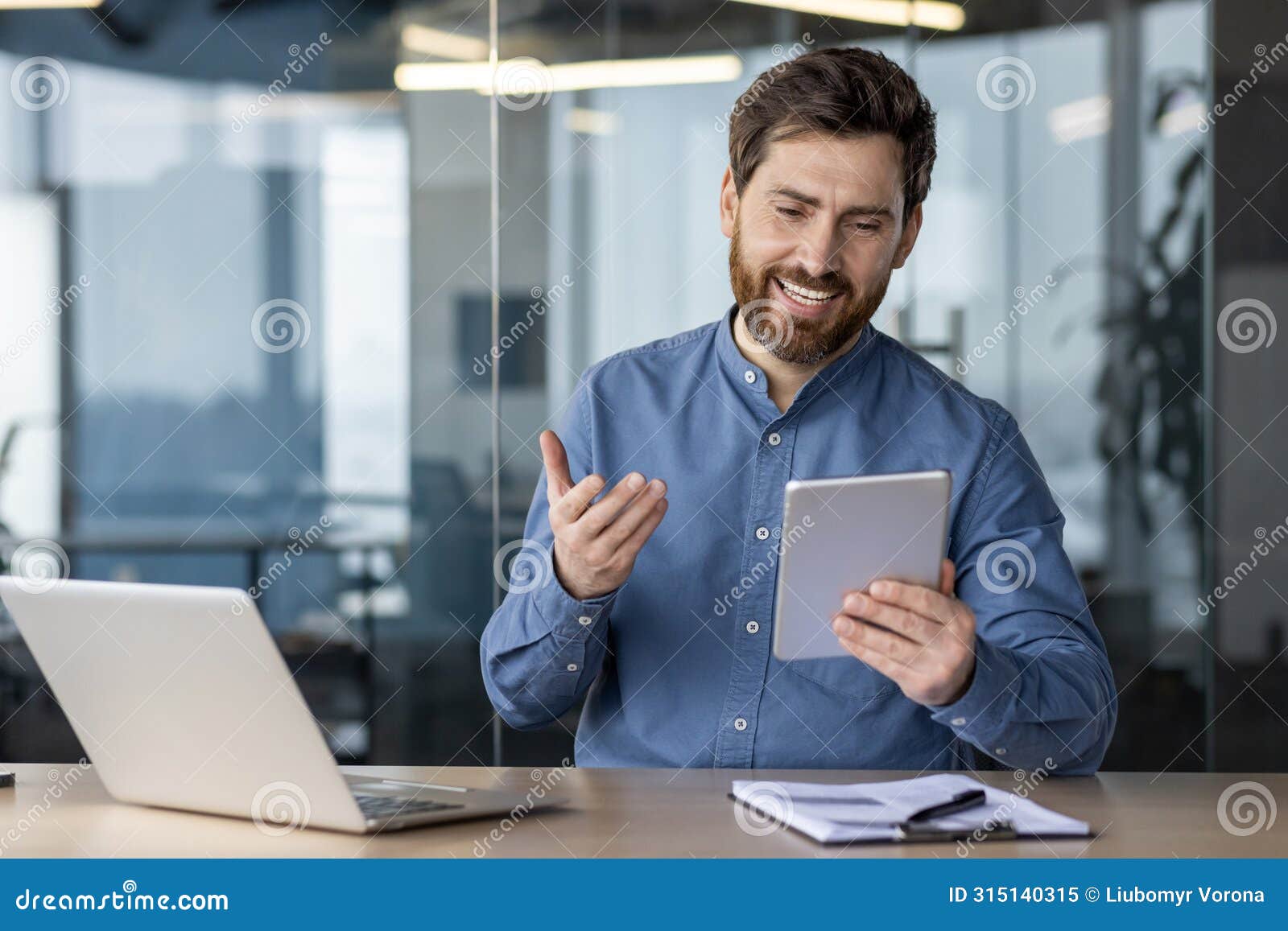 a young man sits in the office at a desk with a laptop and smilingly talks on a video call on a tablet