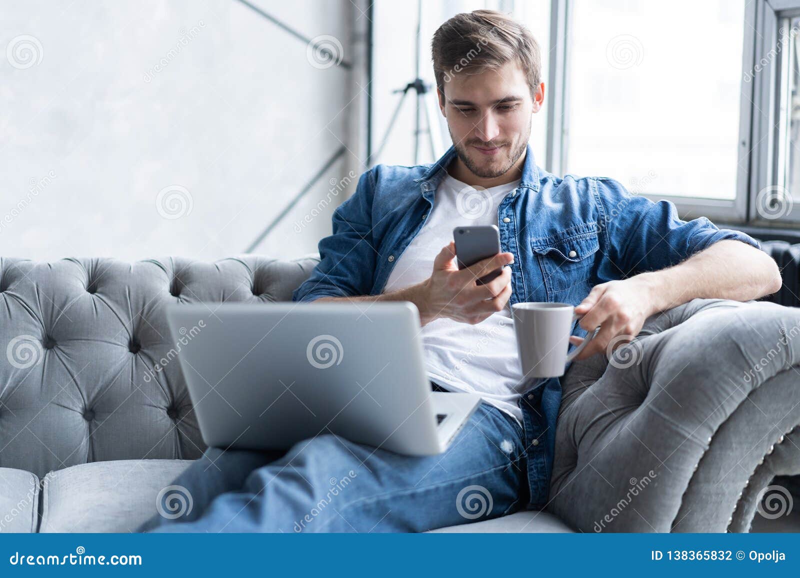 young man using his smartphone for online banking - sitting on sofa with laptop on leap.