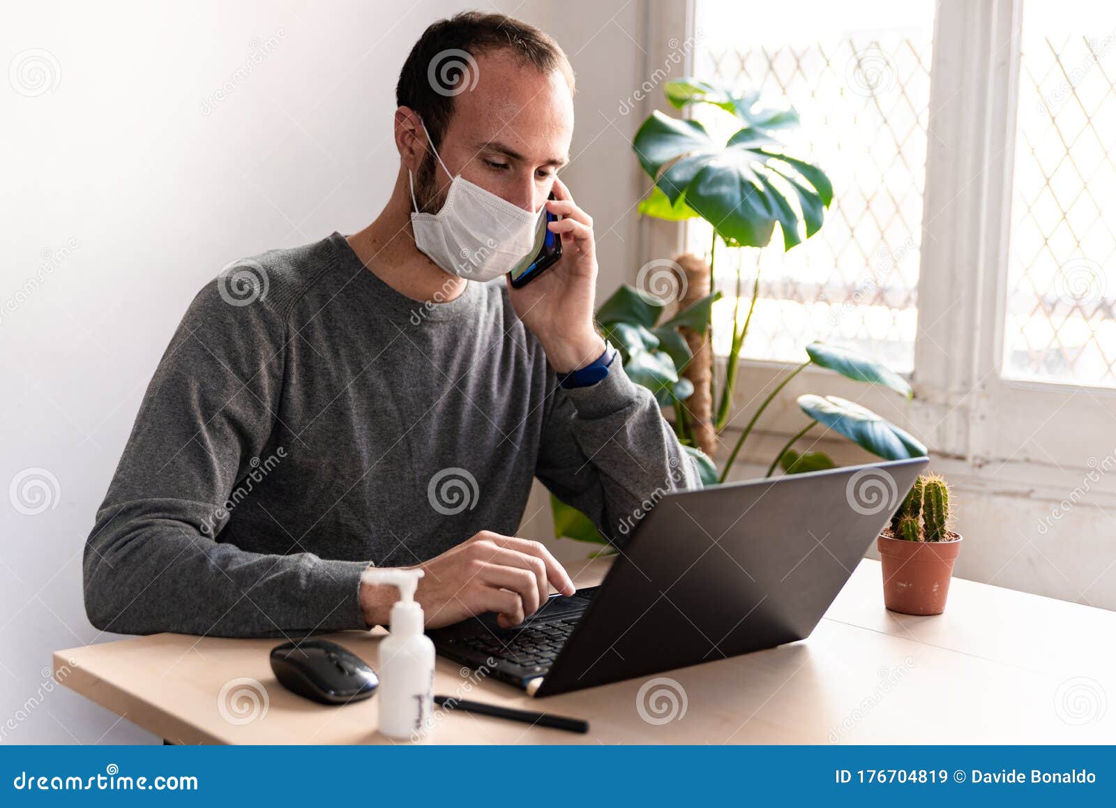 young man wearing medical face mask at work from home due to corona virus outbreak with laptop and mobile phone on wooden table,