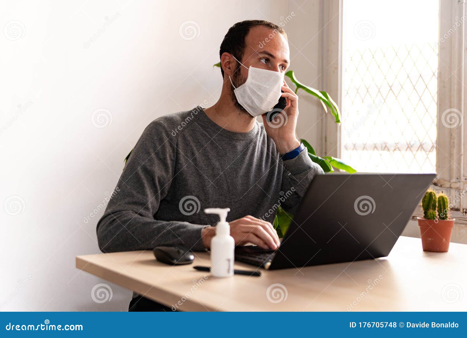 young man wearing medical face mask at work from home due to corona virus outbreak with laptop and mobile phone on wooden table,