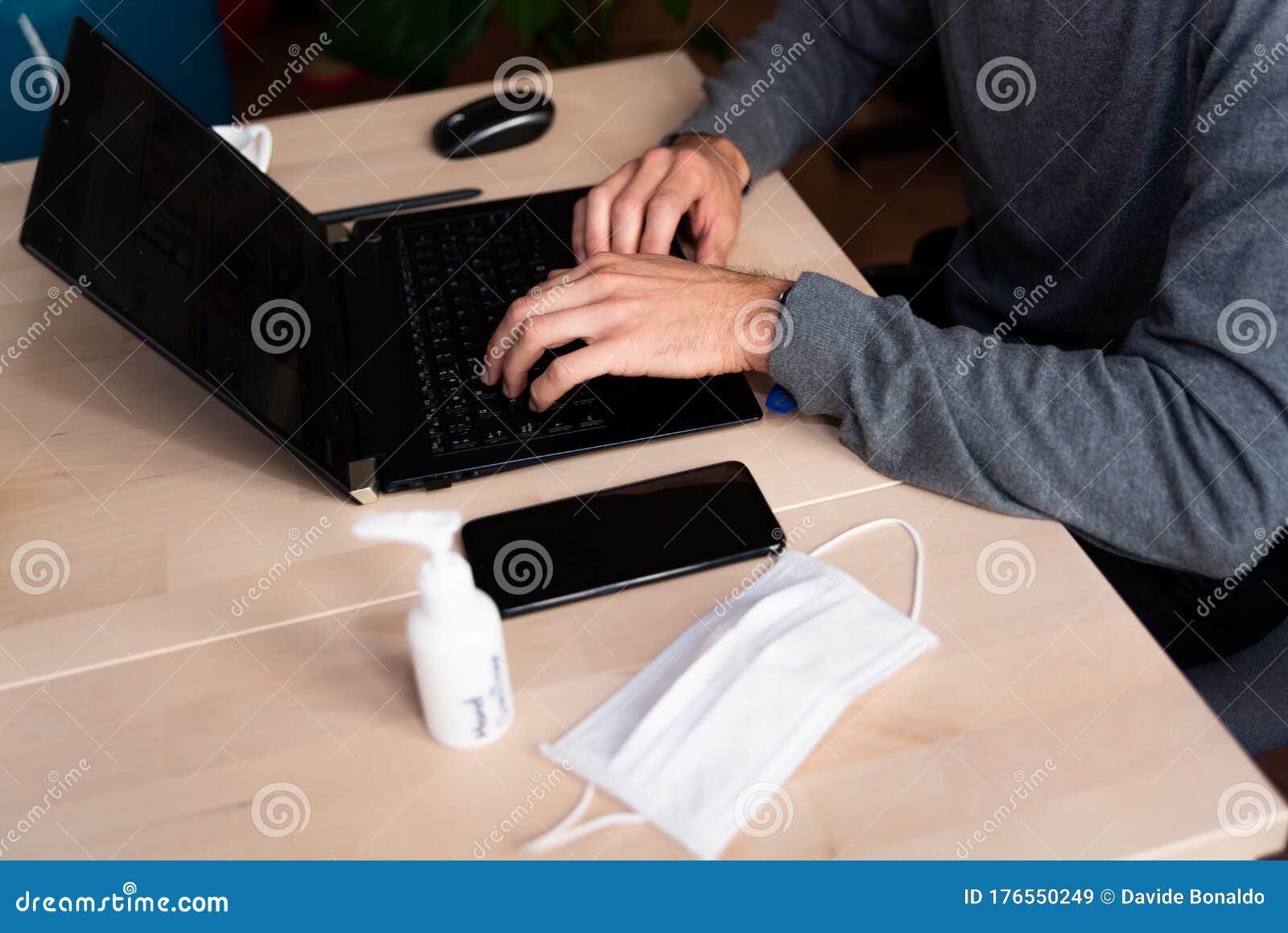 young man wearing medical face mask at work from home due to corona virus outbreak with laptop and mobile phone on wooden table,