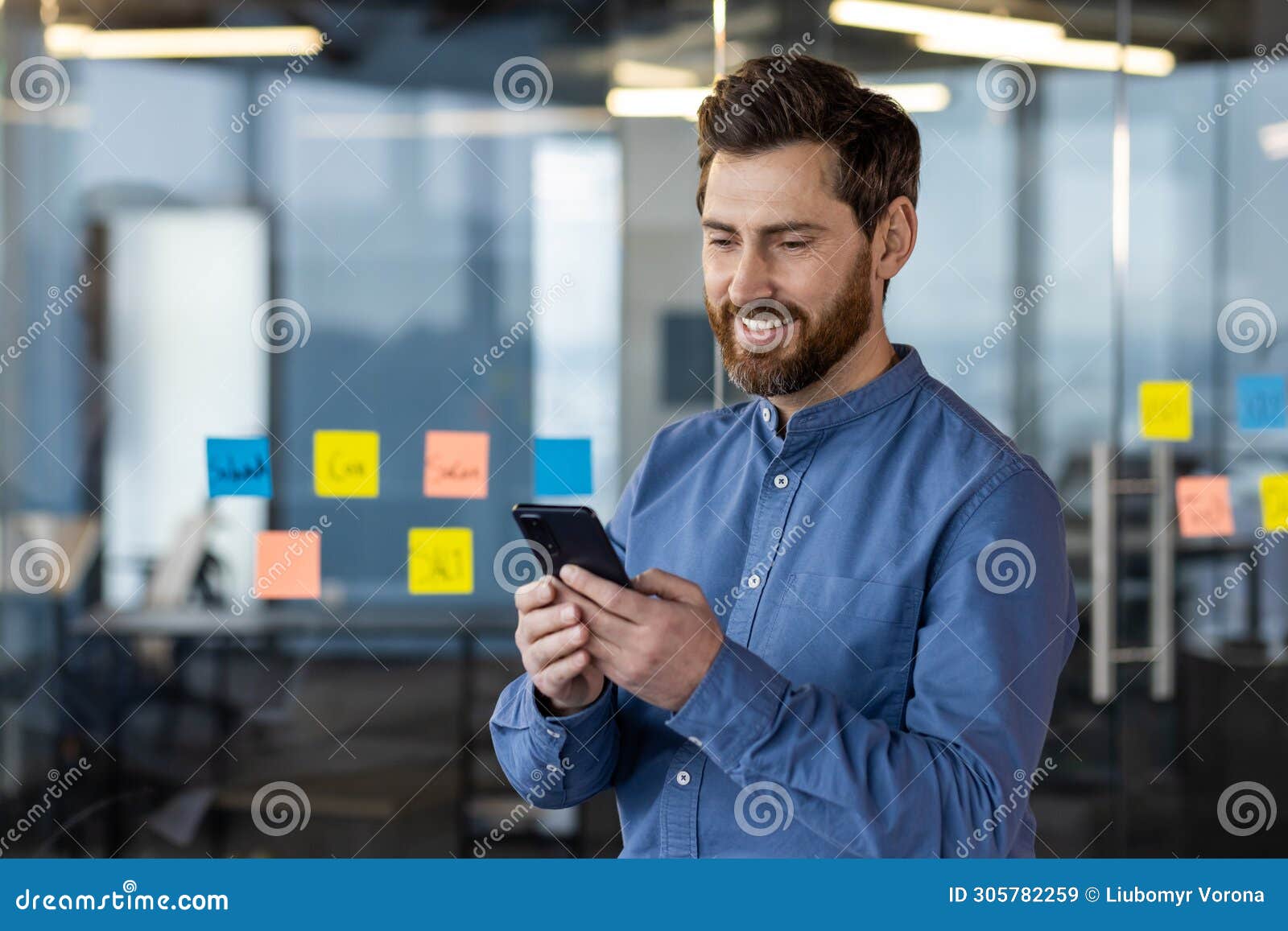a young man is working and standing in the office against the background of a glass board with notes and smilingly using