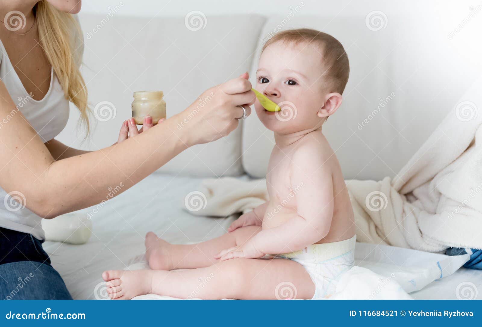 Young Mother Feeding Her Baby Son In Diapers On Sofa At ...