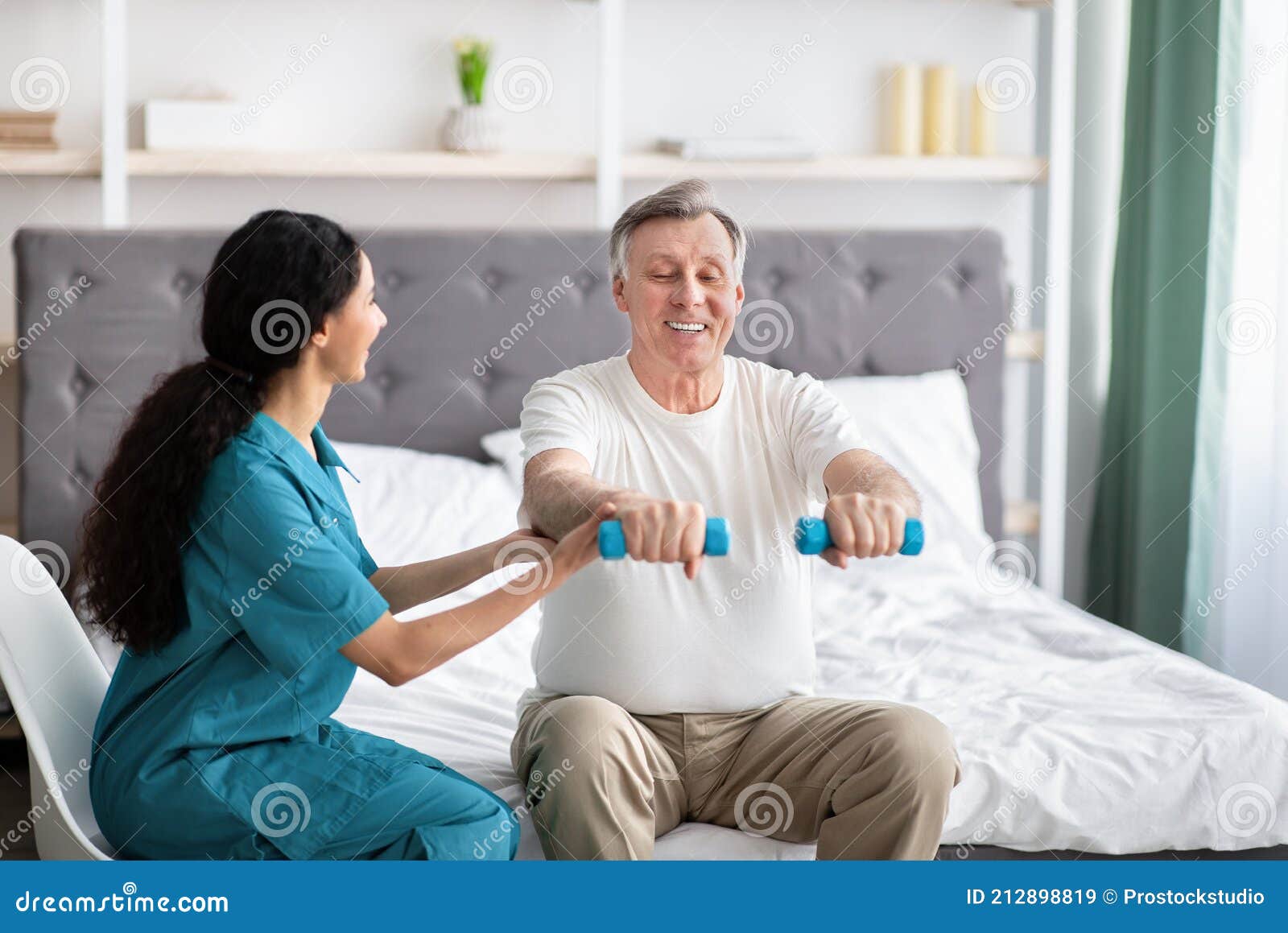 young physiotherapist helping homebound elderly man to do physical exercises with dumbbells on bed at home