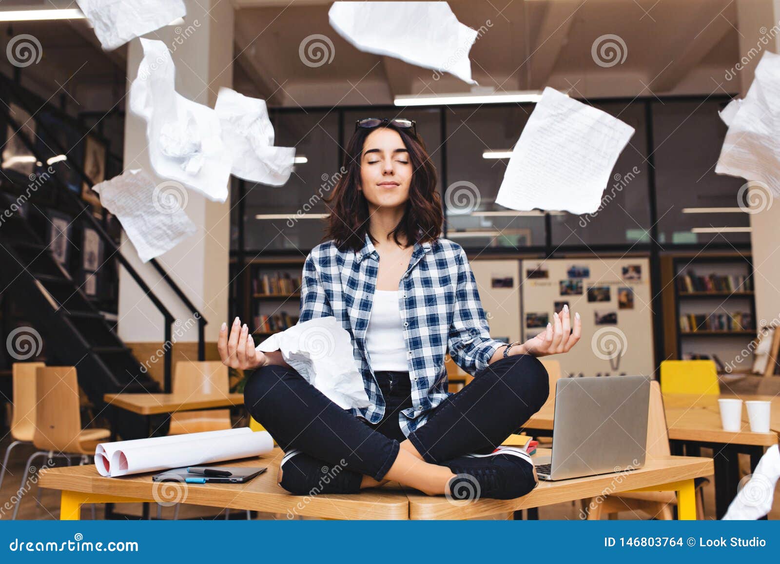 young pretty joyful brunette woman meditating on table surround work stuff and flying papers. cheerful mood, taking a