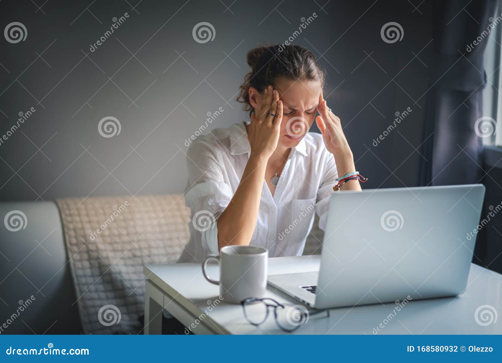 young pretty woman in a white shirt sits at a table in front of a laptop, her eyes and head ache, workaholism, stress burnout