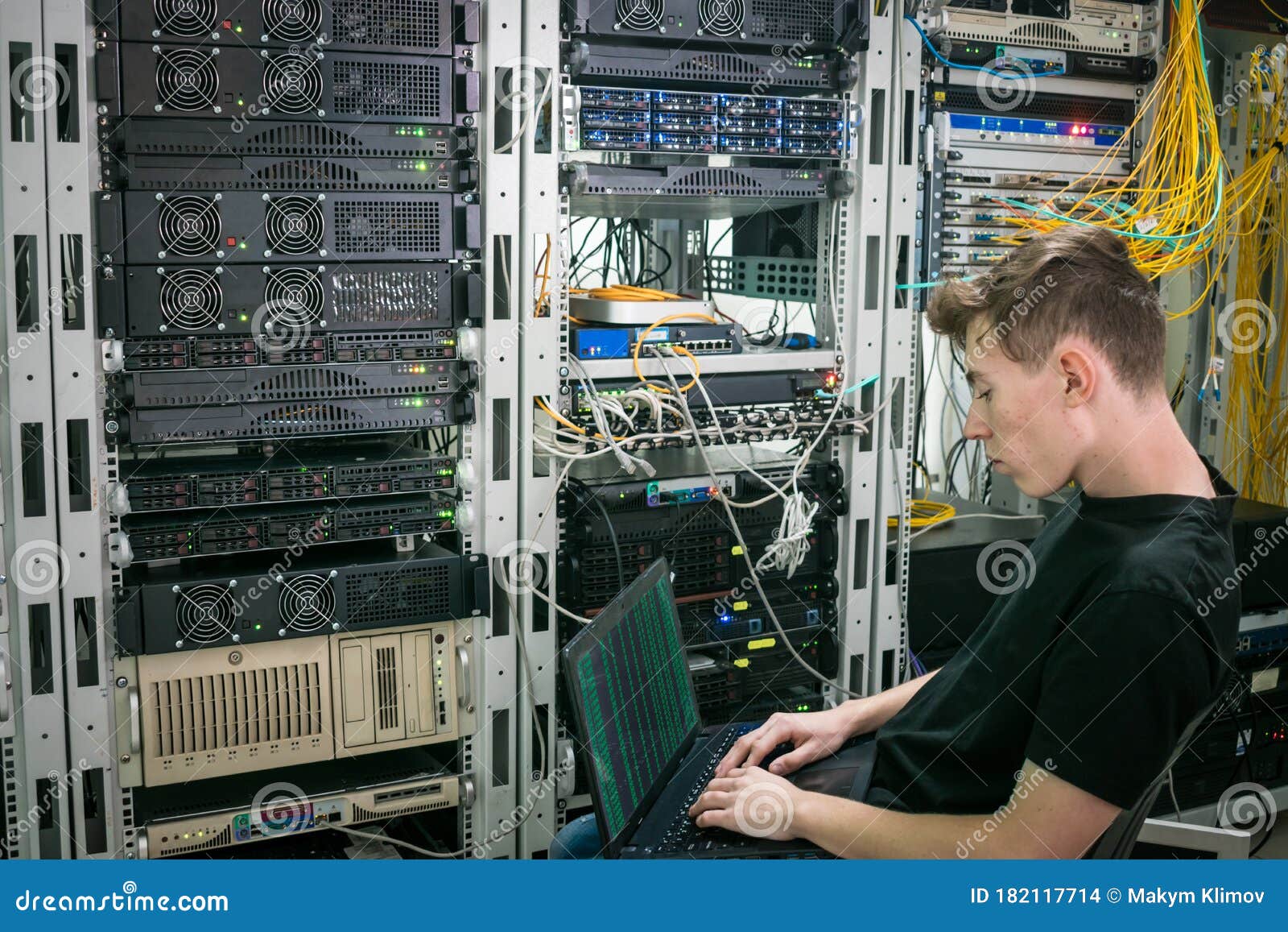 A Young Programmer is Working on a Laptop Near the Racks with Computer