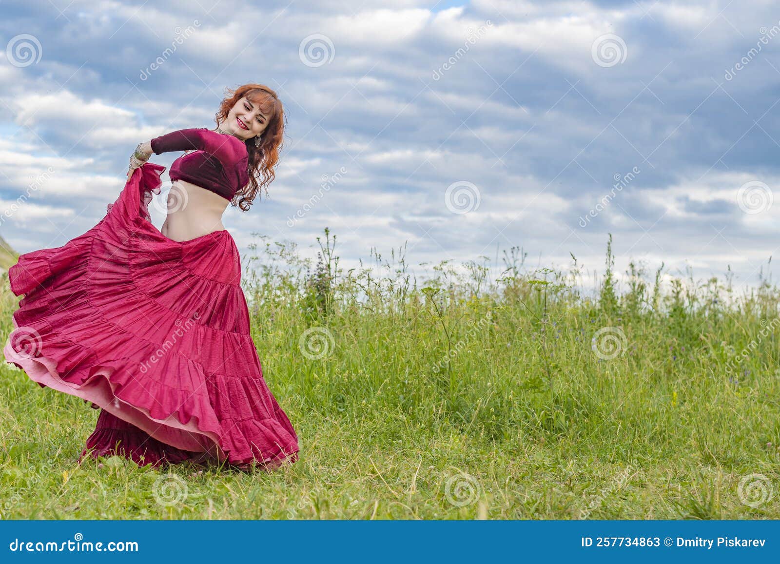 young red-haired girl in red dance dress dances on green meadow against blue summer sky.