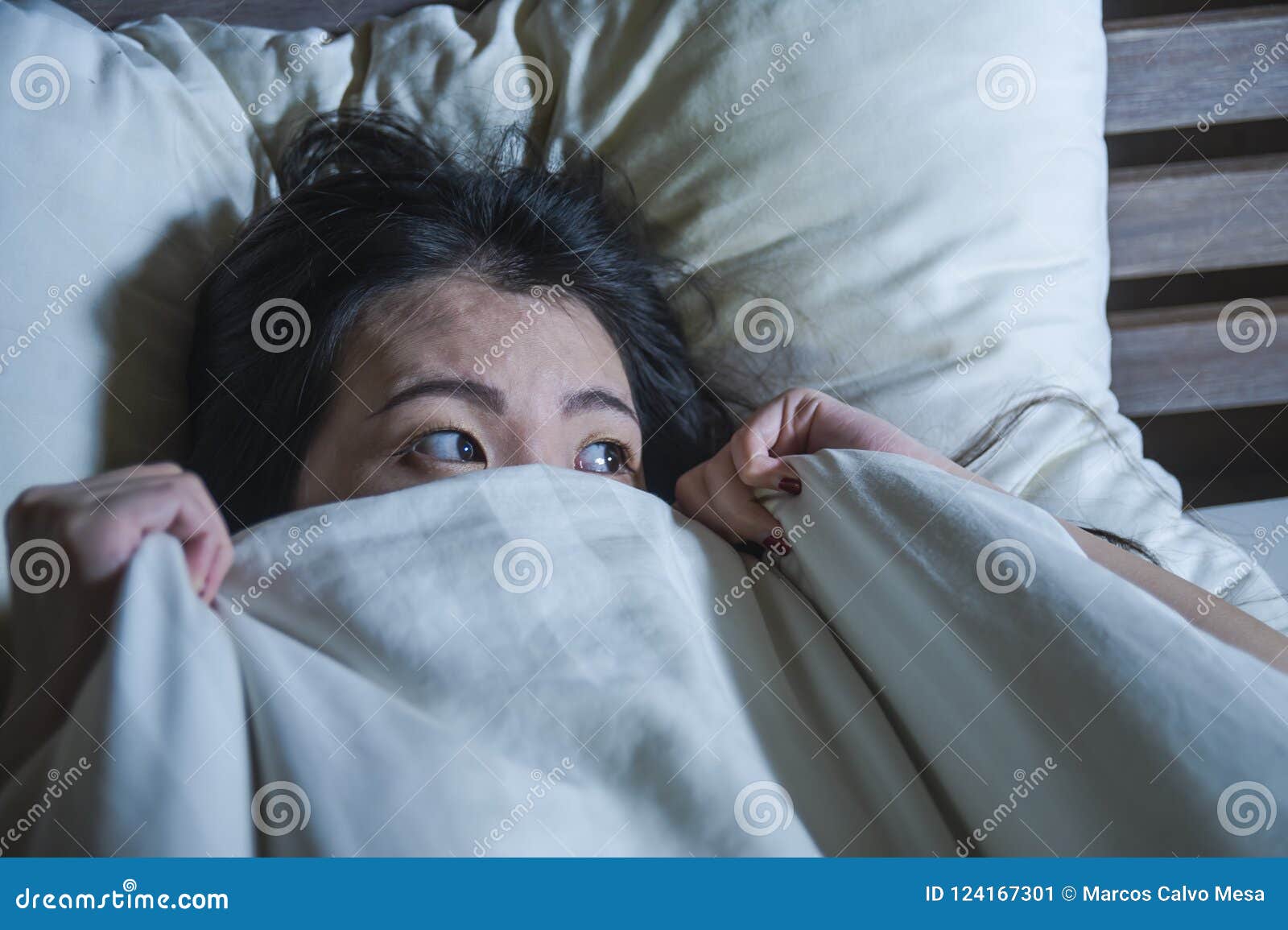 young scared and stressed asian chinese woman lying in bed suffering nightmare in fear and panic grasping blanket covering her hor