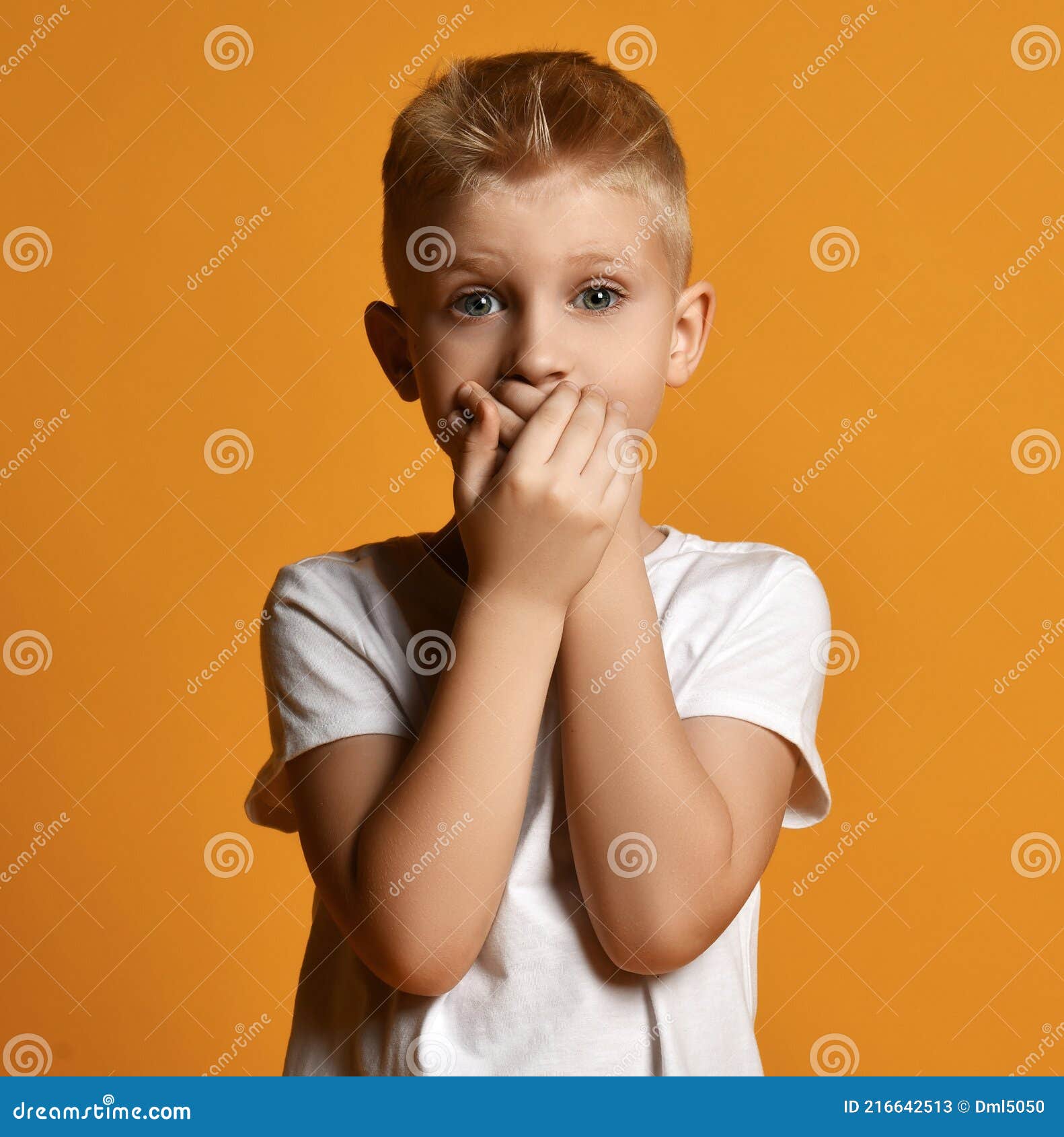young school boy kid in white t-shirt stands scared, eyes wide opened in surprise, covering his mouth with both hands