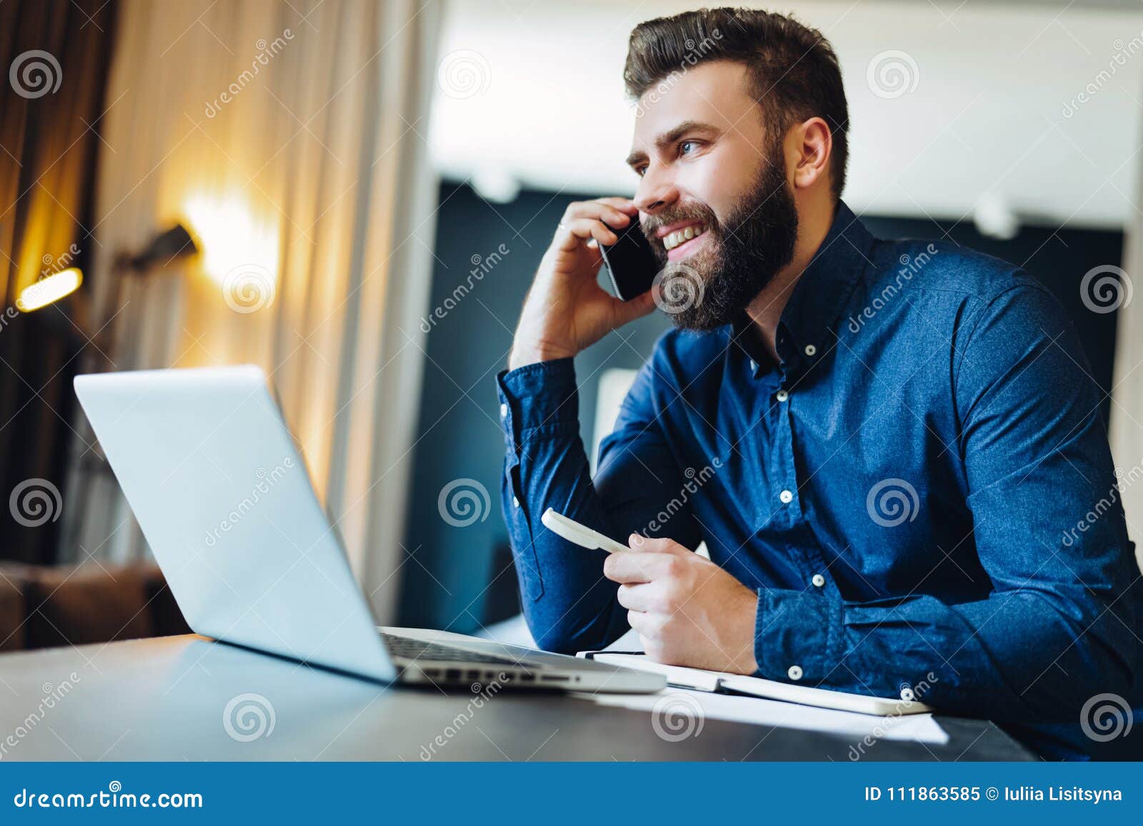young smiling bearded businessman sitting in front of computer, talking on cell phone, holding pen. phone conversations.