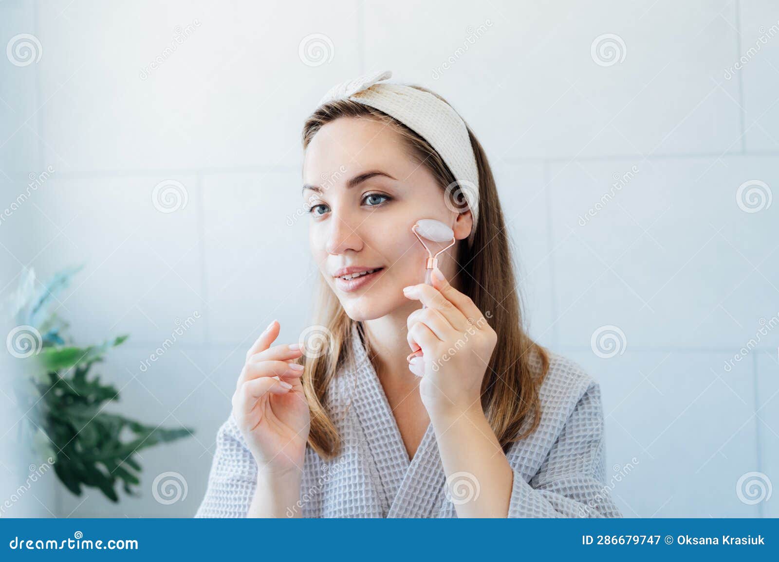 Young Woman In Bathrobe Looking In Mirror And Making Face Massage With Jade Roller In The