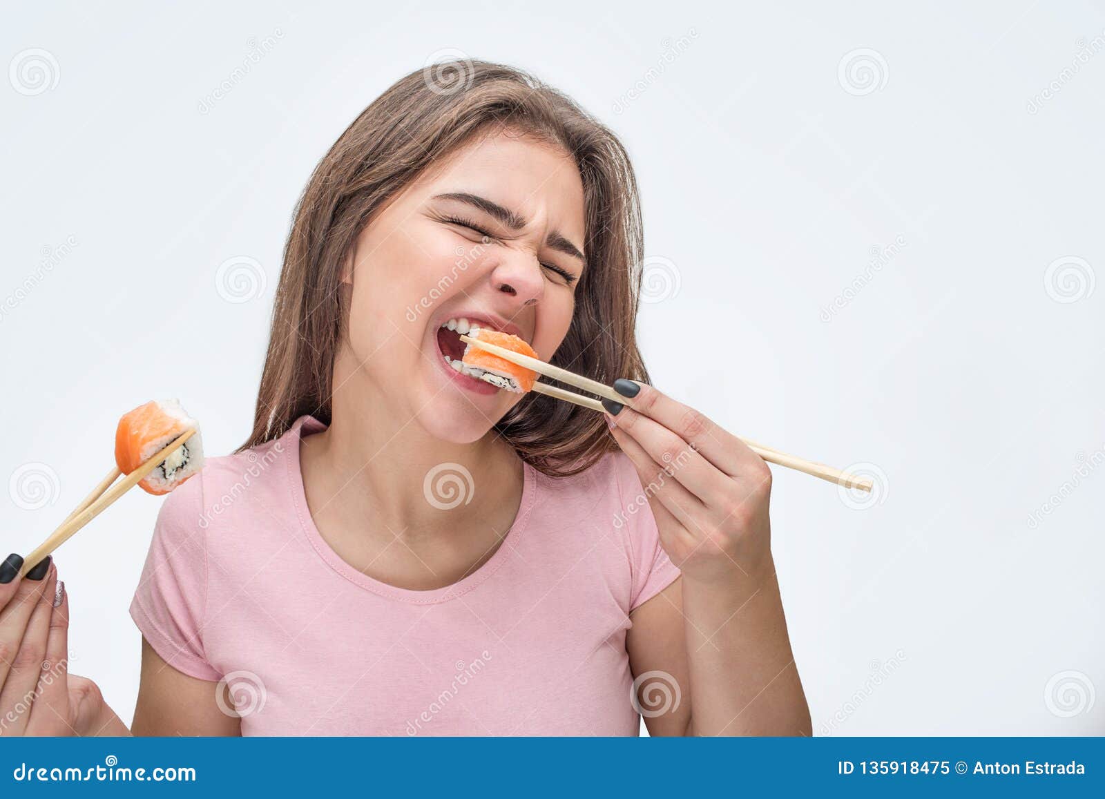 young woman devour pieces of sushi. she bite piece. model hold them with chopsticks.  on grey background.