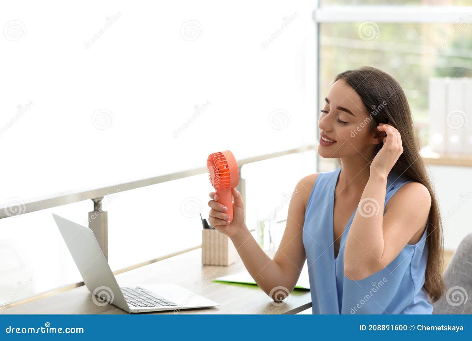 young woman enjoying air flow from portable fan at workplace, space for text. summer heat