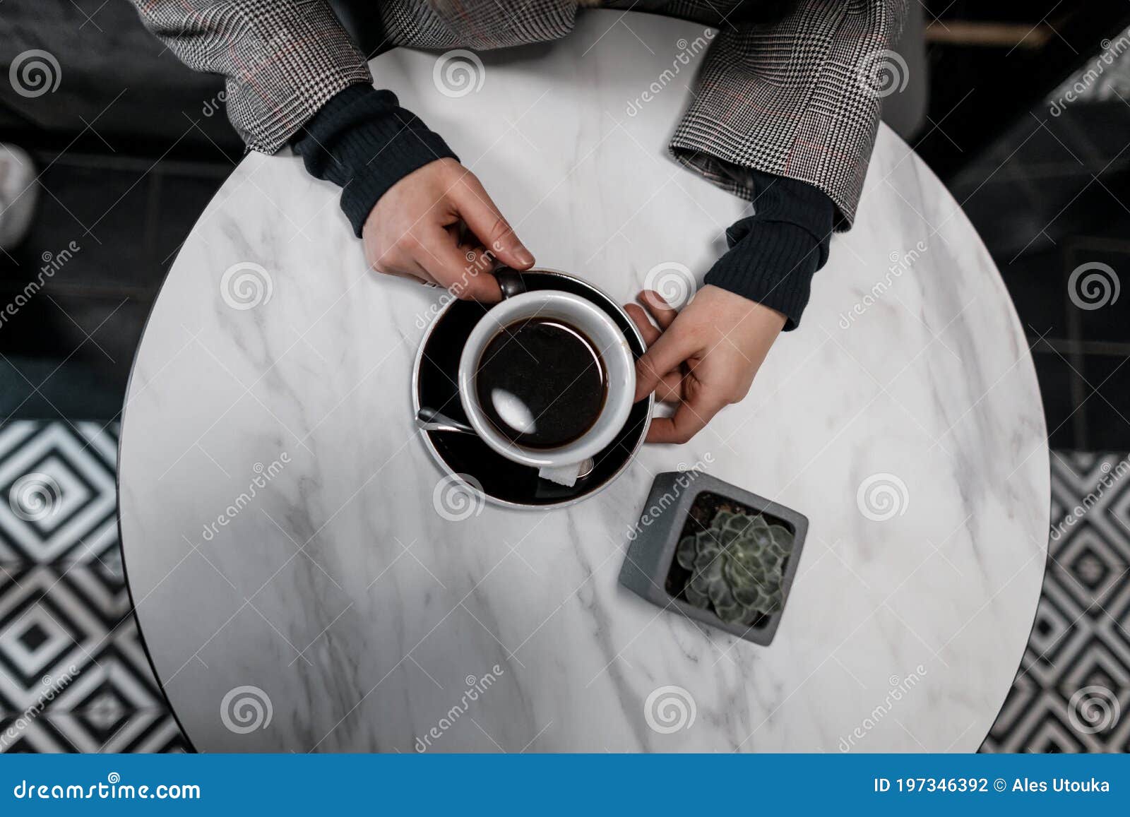 young woman in a fashionable jacket sits at a vintage white table in a cafe and holds in her hand a cup of hot delicious americano