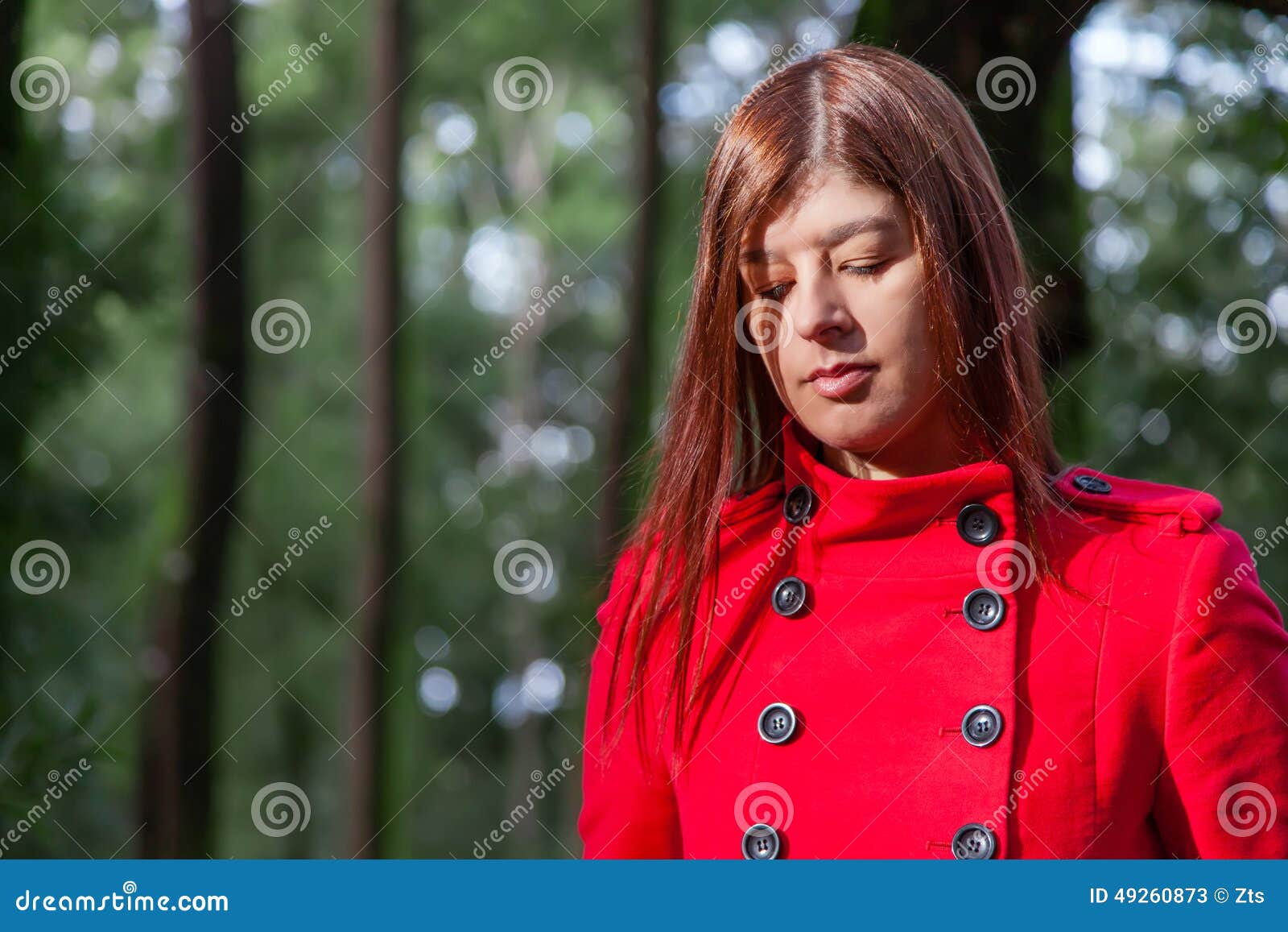 Young woman feeling sad walking on a forest wearing a red overcoat during winter under a sunlight ray