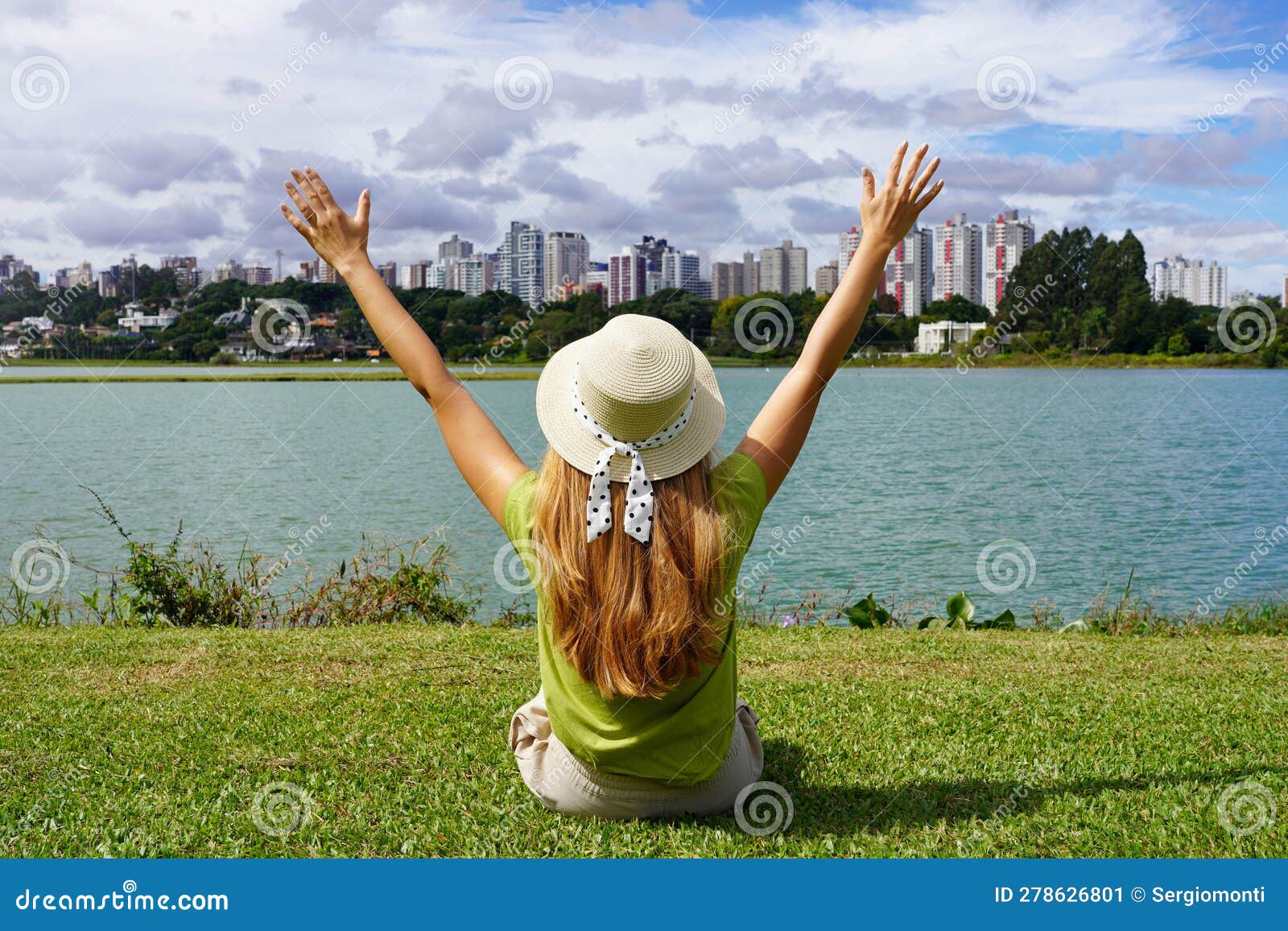 young woman with hat raising arms sitting on grass in barigui park, curitiba, brazil