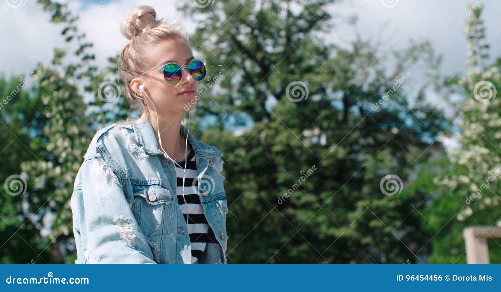 Young woman with headphones enjoying time in a city park. Happy stylish girl wearing denim jacket enjoying time during sunny day. Beautiful girl listening to a music, outdoors.