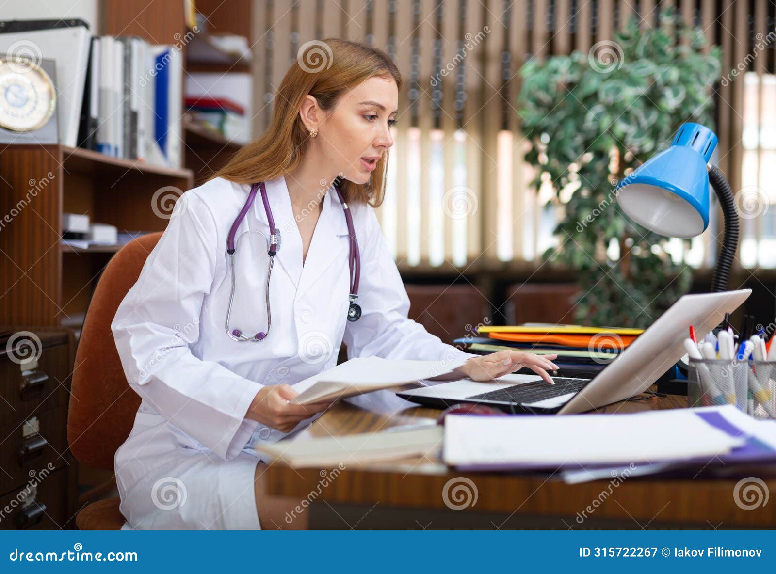 young woman internist studies the patient's outpatient card, typing the treatment appointment on the computer in the