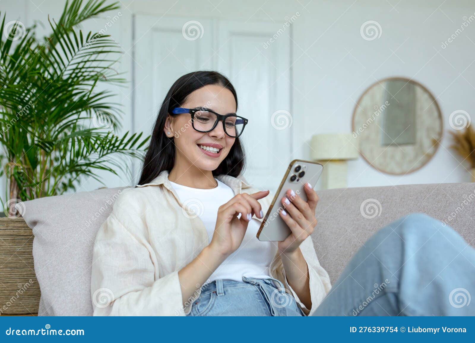 a young woman is lying on the sofa at home and smilingly using a mobile phone
